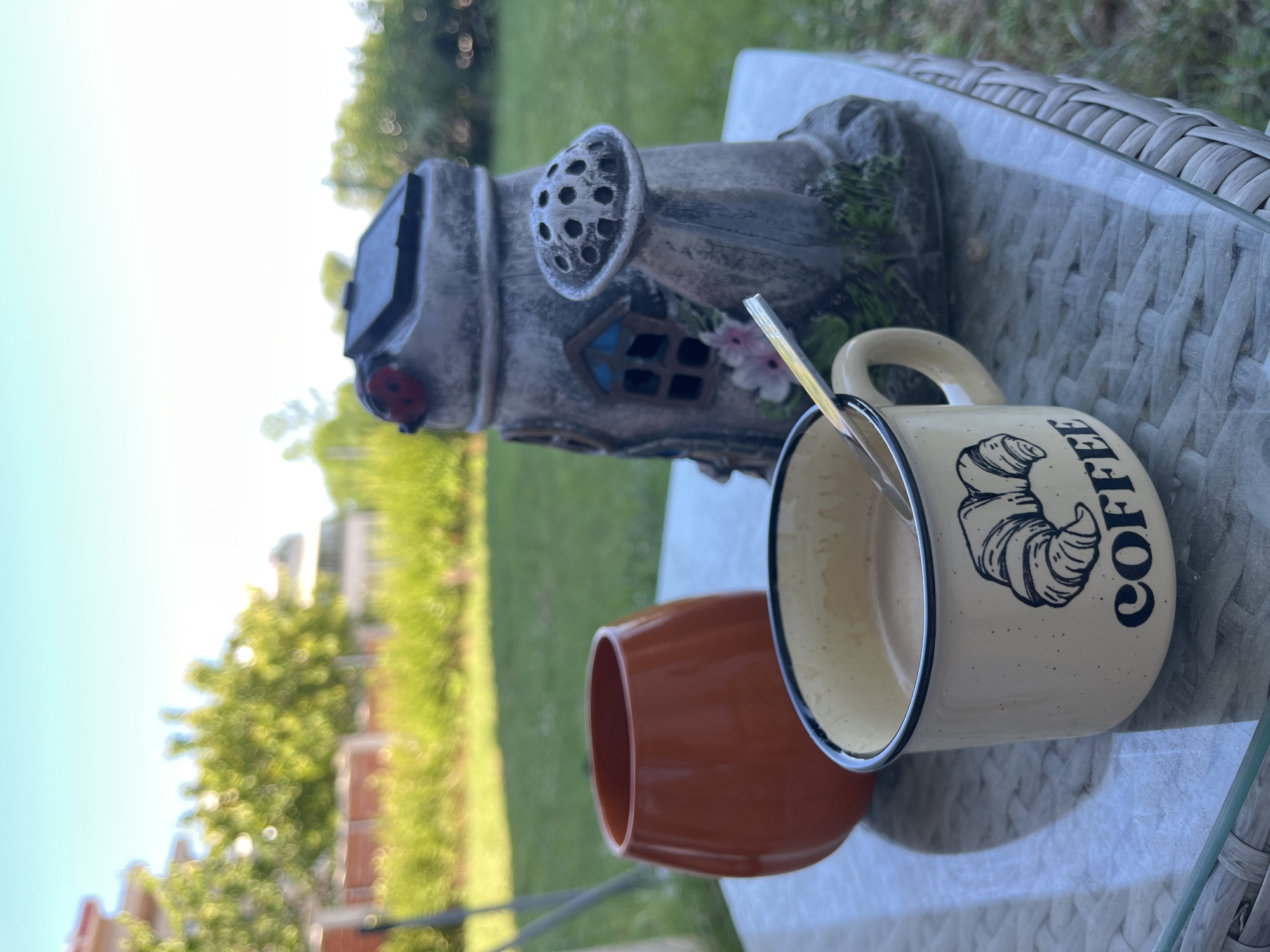 In Tsarevo, Bulgaria, a close-up shot captures a coffee mug with a croissant illustration and the word "COFFEE" emblazoned on it, placed on a glass-top table. A spoon rests inside the mug, which contains a light brown beverage. To the left of the mug is a rounded, orange ceramic vase, and behind them stands a decorative garden gnome shaped like a whimsical house with a ladybug on top and a mushroom-like spout. The background reveals a sunlit garden with green grass, trees, and blurred buildings, suggesting an outdoor setting.
