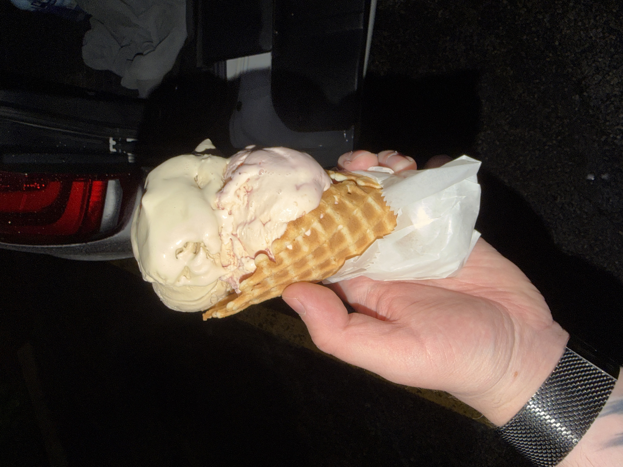 A close-up, low-angle shot shows a hand holding an ice cream cone with two scoops of ice cream. The ice cream is a light beige color and appears to have pieces of strawberry or raspberry mixed in. The cone is a golden brown waffle cone, wrapped in white paper. The hand is holding the cone from the bottom, with fingers wrapped around the paper. The background is dark, with a blurry image of a vehicle's taillight and the open trunk of a car. The ground appears to be asphalt. The scene is set in the city of Arlington Heights, United States of America.