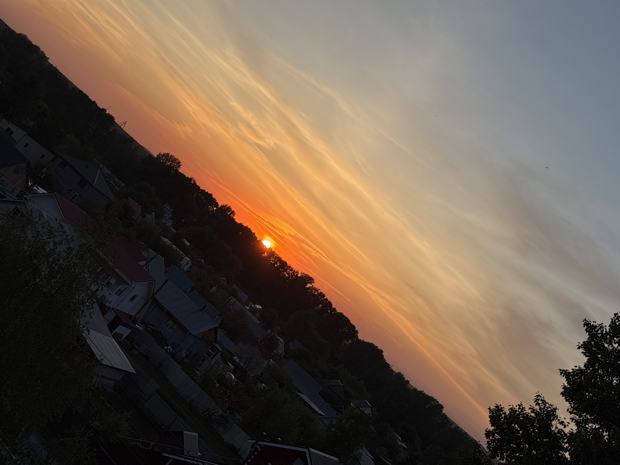 The image shows a sunset in a residential area of Chaikovsky, Russia. The sun is setting behind trees and houses, casting an orange glow across the sky. The houses are mostly dark, and the trees are silhouetted against the sky. The image is taken from an elevated position, giving a view of the rooftops and trees.