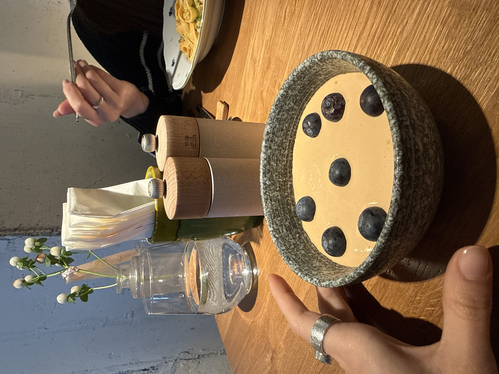 The image depicts a table scene, likely within a restaurant or dining establishment. A person with a ring on their finger is seen in the lower right corner near a bowl. The bowl contains a pale yellow substance, possibly a dessert or pudding, dotted with blueberries. Adjacent to the bowl are two cylindrical objects, presumably salt and pepper grinders. A clear glass vase, holding a small bouquet of white flowers, and a napkin dispenser are positioned near the grinders. In the upper left corner, a person with a striped garment, holding a utensil, is partially visible, dining at the same table. A plate of pasta is seen in the upper left. The table is made of wood and the background suggests an indoor setting. There is no clear indication this photo was taken in Kazan, Russia.