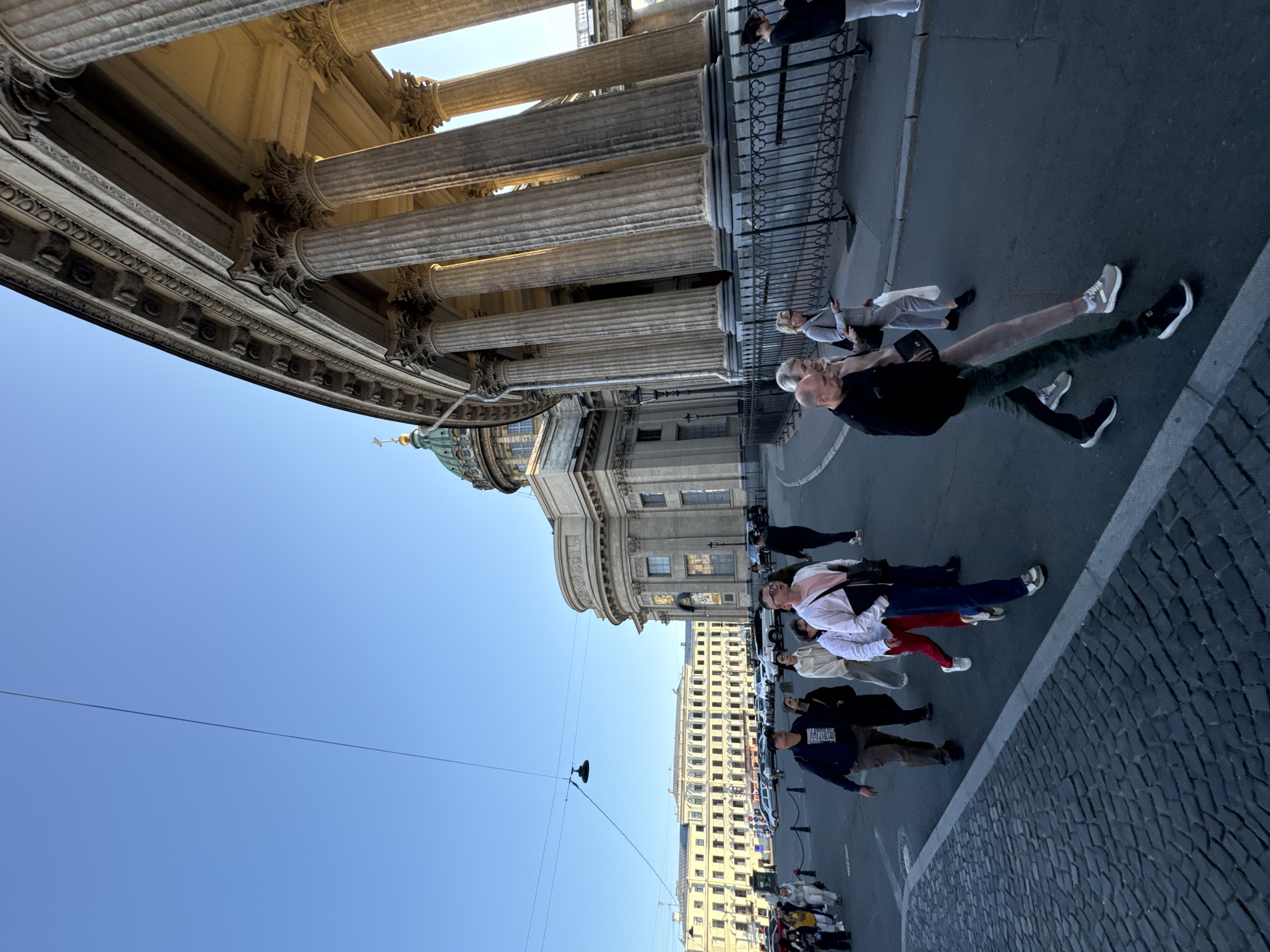 The image depicts the Kazan Cathedral, a prominent landmark in Saint Petersburg, Russia, bathed in the warm light of what appears to be late afternoon or early evening. A wide, paved plaza stretches out in front of the cathedral, with a cobblestone section in the foreground. Several people are walking across the plaza, some in groups, others individually.
The cathedral itself is grand, with a semicircular colonnade of massive Corinthian columns dominating the right side of the frame. Behind the colonnade, the main body of the cathedral rises, featuring a large, green dome topped with a golden cross. The architecture is classical and imposing.
In the middle ground, a long, pale yellow building with numerous windows lines the edge of the plaza, contrasting with the monumental stone of the cathedral. Several cars are parked or driving in this area, indicating it is a public street and thoroughfare.
The sky is a clear, bright blue, with a single overhead wire and a street lamp visible. The overall impression is of a vibrant, active urban space with a rich architectural heritage. The people in the image appear to be tourists or locals going about their day, their presence adding a sense of scale and life to the majestic surroundings.
