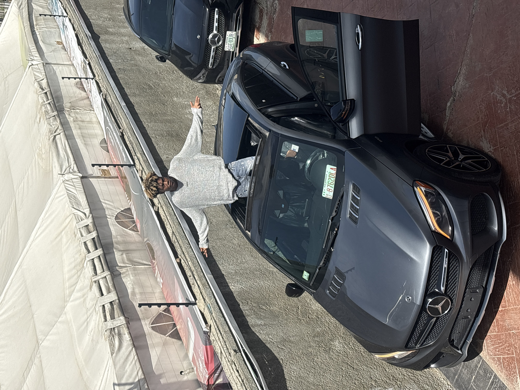 The image depicts a man in a light gray sweater and denim jeans leaning out of the driver's side window of a dark gray Mercedes-Benz coupe. The car has a license plate that reads "8762DLA". The car's driver's side door is open. Another dark-colored Mercedes-Benz is parked behind it with a license plate that reads "G6E 03JB". A large white tent is positioned on the left side of the frame. The scene is set in Igboefon, Nigeria, and the ground appears to be a mix of paved and brick surfaces.