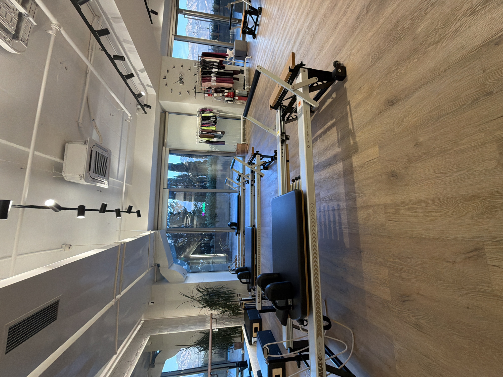 The image depicts the interior of a modern Pilates studio located in Boreti, Montenegro. The room is spacious with light wood flooring and large windows offering a view of trees and mountains. Several Pilates reformers, specifically Merrithew V2 Max reformers, are neatly arranged in the foreground and middle ground. These machines are predominantly white and black with wooden accents on the footbars and seats. In the background, there are clothing racks displaying workout attire, a wall clock with a bird design, and potted plants. The lighting is a mix of natural light from the windows and overhead track lighting. No people are visible in the scene.