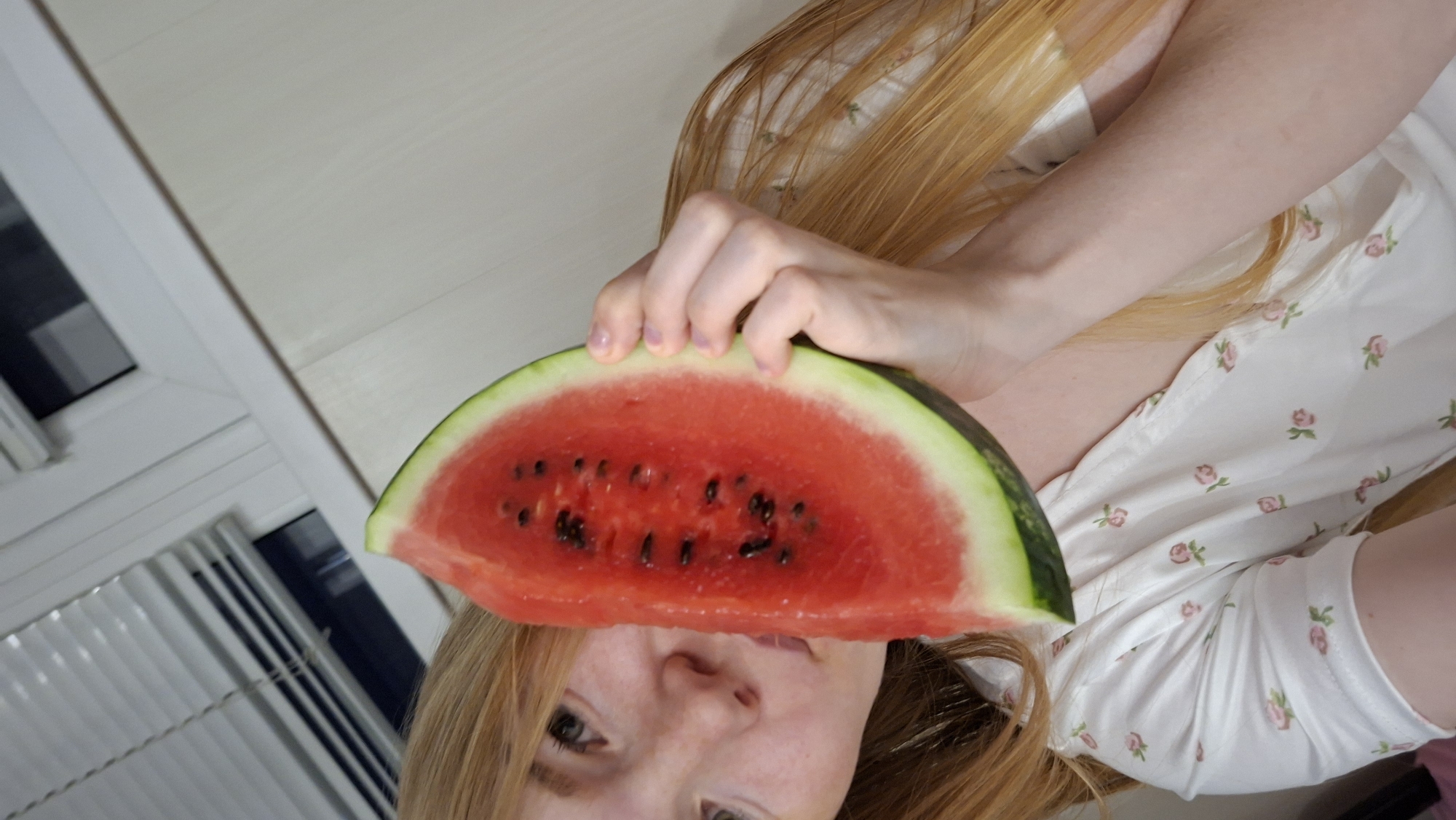 A young woman with long blonde hair is holding a slice of watermelon up to her face, partially obscuring it. She is wearing a white blouse with a pink floral pattern. The background appears to be an indoor setting, possibly a room, with a window and blinds visible. The overall lighting is soft and natural. There are no specific landmarks or details to suggest the location is in Chaikovsky, Russia.