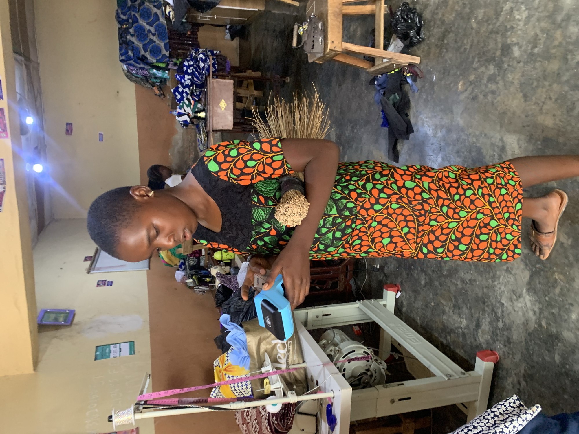 The image captures an indoor scene within a bustling tailoring or craft workshop in Jos, Nigeria, during the daytime. The main subject is a young person, likely female, with short, dark hair, wearing a vibrant ankle-length dress. The dress features an eye-catching pattern of orange and green leaf-like shapes on a black background, with a plain black bodice. She stands, looking downwards with a focused expression, engaged in an activity.
In her right hand, she holds a light blue tool, possibly a tagging gun or label applicator, which she is operating over a piece of light-colored fabric or an item laid out before her. Her left arm is bent, and she holds a bundle of thin, dried sticks, resembling small whisk brooms, against her hip.
The immediate foreground reveals parts of a white sewing machine stand, displaying the brand name "EMEL." A pink measuring tape, with visible markings like "31, 32, 33," is draped over the stand, alongside spools of thread and various fabric scraps. The walls of the room are painted in two horizontal sections: a lighter, cream-colored upper portion and a brown lower section. Bright reflections from light sources are visible on the upper wall. Faint white writing, resembling chalk, is present on the wall, and a green rectangular sign can be seen further back.
The background and surrounding areas are filled with materials and equipment typical of a workshop. Piles of patterned fabrics, some folded, others draped, are visible on wooden shelving and structures. A larger bundle of the dried sticks, similar to those held by the subject, rests on a surface in the mid-ground. The floor is a dark, unpolished concrete. The overall impression is one of an active and functional workspace.
