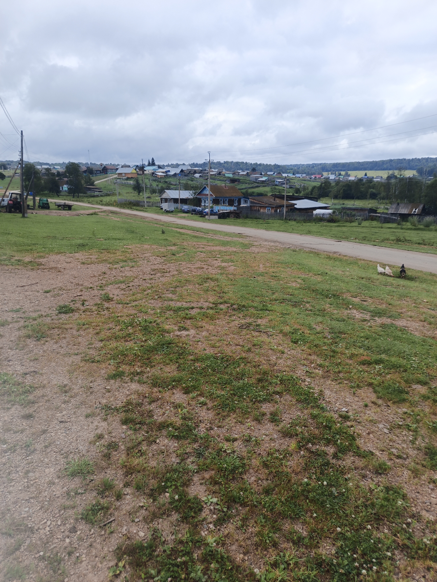The image depicts a rural landscape in Islambaevo, Russia, under an overcast sky. In the foreground, a dirt and gravel path transitions into a grassy area with sparse vegetation. A dirt road runs horizontally across the midground, separating the foreground from a village scene. The village consists of several wooden houses, some with blue trim, and other structures, appearing to be residential and possibly agricultural buildings. A few vehicles, including a tractor and a trailer, are visible on the left side of the road near the village entrance. In the distance, the village extends further, with more houses and buildings nestled amongst trees. The background features a line of dense green trees and hills, suggesting a natural, undeveloped area surrounding the settlement. Three chickens, two white and one dark, are present in the grassy area near the road in the mid-right portion of the image. Power lines and utility poles are also visible throughout the scene, indicating the presence of infrastructure. The overall impression is one of a quiet, rural community in a natural setting.