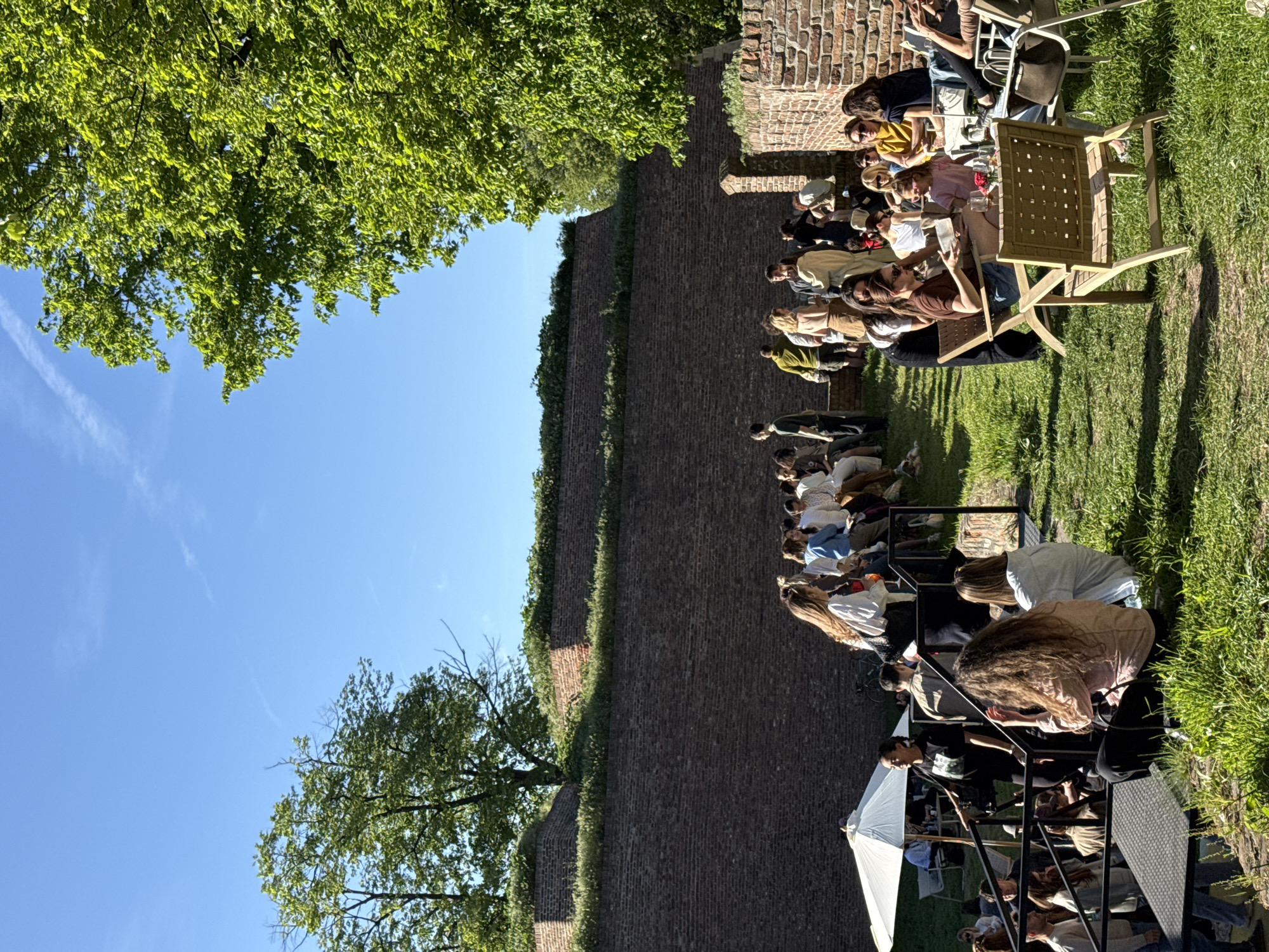 A sunlit outdoor scene shows a group of people gathered along a brick wall in Belgrade, Serbia. The brick wall runs vertically, separating the scene into left and right halves. On the left, a large tree and clear blue sky are visible. On the right side of the wall, people sit on chairs and benches along the grass. Some people are seated at tables, suggesting a social gathering, possibly a cafe or outdoor event. An umbrella is visible among the people. The scene indicates a relaxed, warm atmosphere.