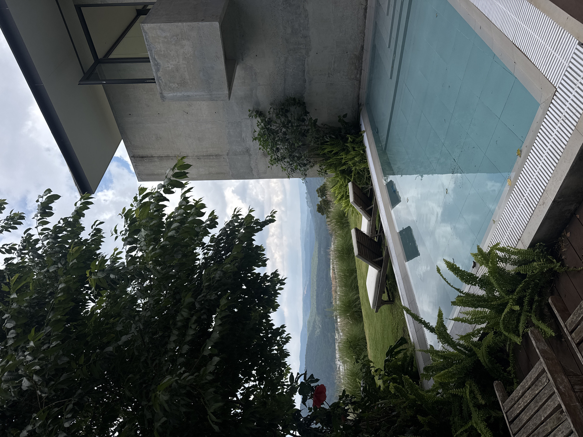 This image depicts a serene outdoor scene with a modern architectural element overlooking a lush landscape. The foreground features a swimming pool with clear, light blue water, reflecting the sky and surrounding greenery. The pool's edge is lined with a light-colored coping and a white grated drain. To the left of the pool, weathered wooden chairs are visible on a wooden deck.
Beyond the pool, a well-maintained green lawn is dotted with tall, wispy grasses. Two empty chaise lounges with light-colored cushions are positioned on the lawn, facing the distant view. In the mid-ground, a dense canopy of dark green leaves from a large tree overhangs the scene, with a single bright red hibiscus flower visible on its left side.
The background reveals a rolling landscape of hills covered in dense vegetation, fading into a hazy blue in the distance under a cloudy sky.
To the right of the pool, a striking modern structure made of concrete dominates the view. This building features clean lines, with a cantilevered section and large glass windows. A small green bush and ferns grow against the concrete wall near the pool.
While the image is labeled as being in Đa Lộc, Vietnam, there are no people visible in the scene, and no specific landmarks identifying it as being within a city. The overall impression is one of tranquility and luxurious seclusion, likely a private residence or resort.
