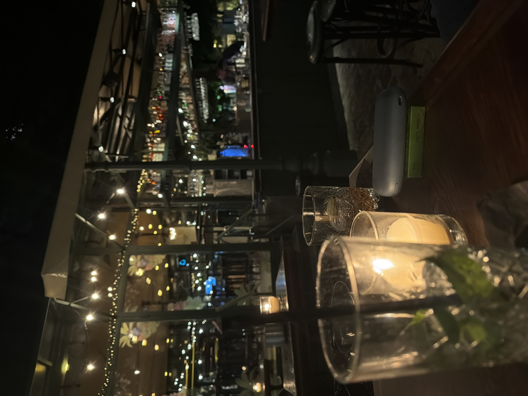 This nighttime photo shows a bar setting, likely in Sofia, Bulgaria. In the foreground, two glasses are visible on a dark wooden surface. One contains a sprig of greenery, possibly mint, while the other holds a beverage with a straw. A small cylindrical electronic device and a green package rest on the table near the glasses. The background features a blurred view of a bar with bottles lining shelves, illuminated by lights. String lights are draped around what appears to be a glass-enclosed structure, possibly an outdoor patio or garden area of the bar. The overall lighting is dim, with a warm glow emanating from the candles and string lights, creating a cozy atmosphere.