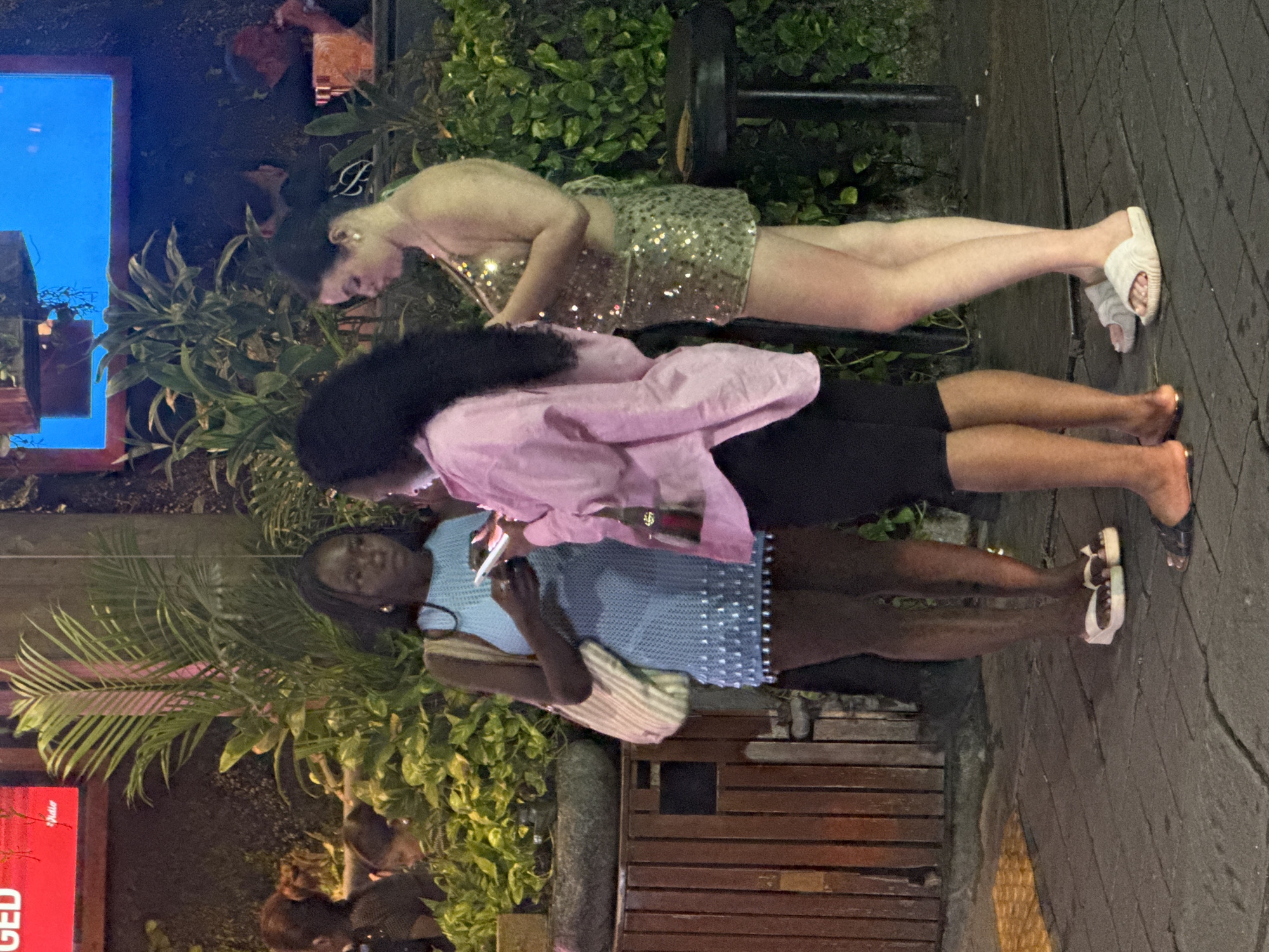Three individuals, two women prominently in the foreground and a third less visible person in the background, are standing on a paved walkway at night. The woman on the left, with dark skin, wears a light blue, textured dress with beaded embellishments and carries a striped tote bag. She is looking down at her phone. To her right, a woman with long dark hair wears a pastel pink oversized shirt and black shorts. She is also looking at her phone. On the far right, a woman with fair skin wears a sparkling gold halter top and a matching sequined skirt. She is looking to her left and slightly down. Behind them, lush green foliage is visible, along with wooden fencing. Overhead, brightly colored neon signs, one red and one blue, are illuminated. The scene appears to be an outdoor establishment in North Kuta, Indonesia, possibly a restaurant or bar, given the ambiance and lighting.
