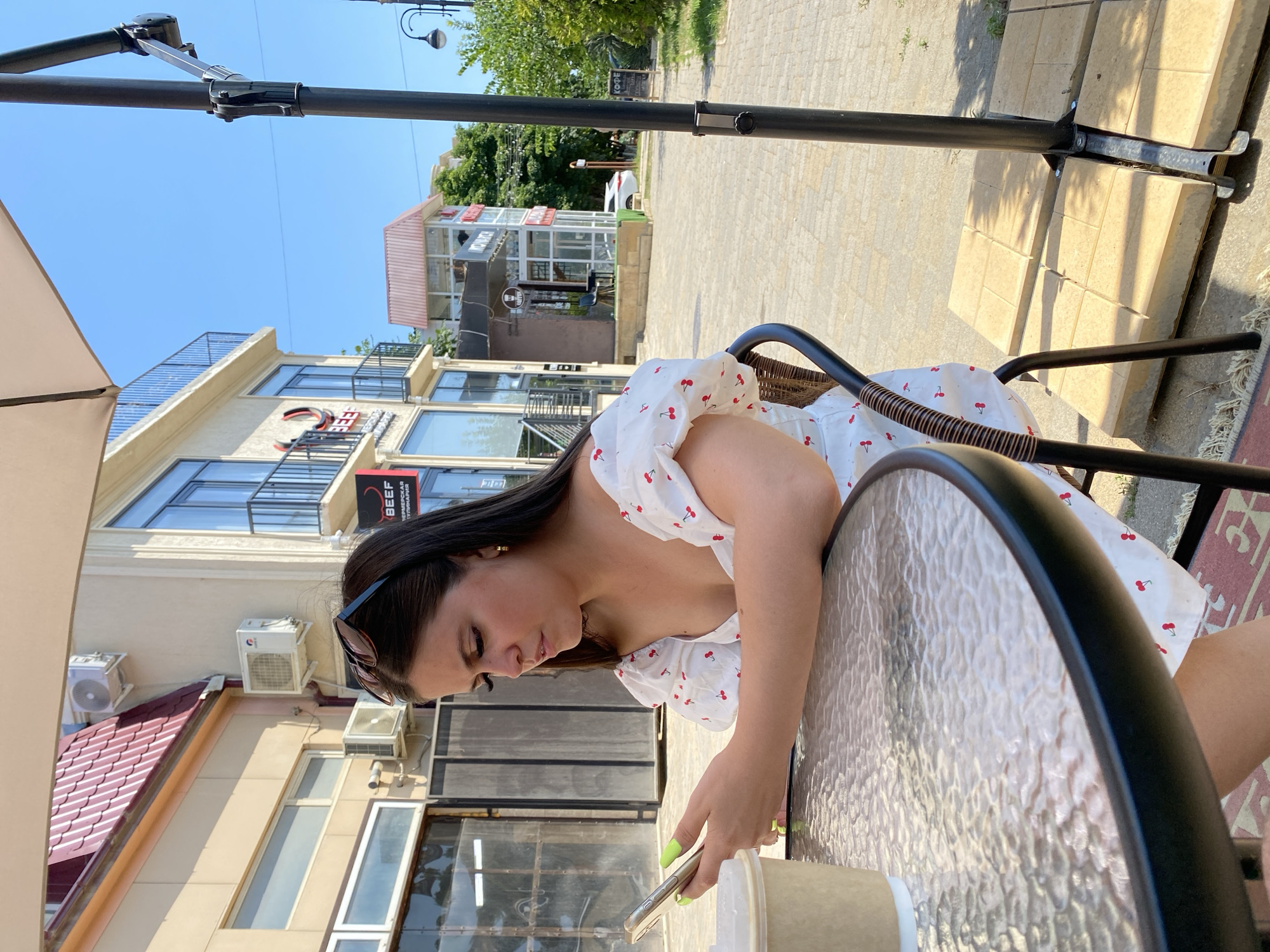 An outdoor cafe scene in Derbent, Russia, features a young woman seated at a glass-topped table, engrossed in her smartphone. She has dark hair, wears a white dress with red cherry patterns, and has bright green fingernails. A disposable coffee cup is on the table in front of her. The cafe appears to be located on a street with a paved walkway and several buildings in the background, including a multi-story structure with balconies and air conditioning units. A large umbrella provides shade over the seating area. The sky is clear and blue, indicating a sunny day.