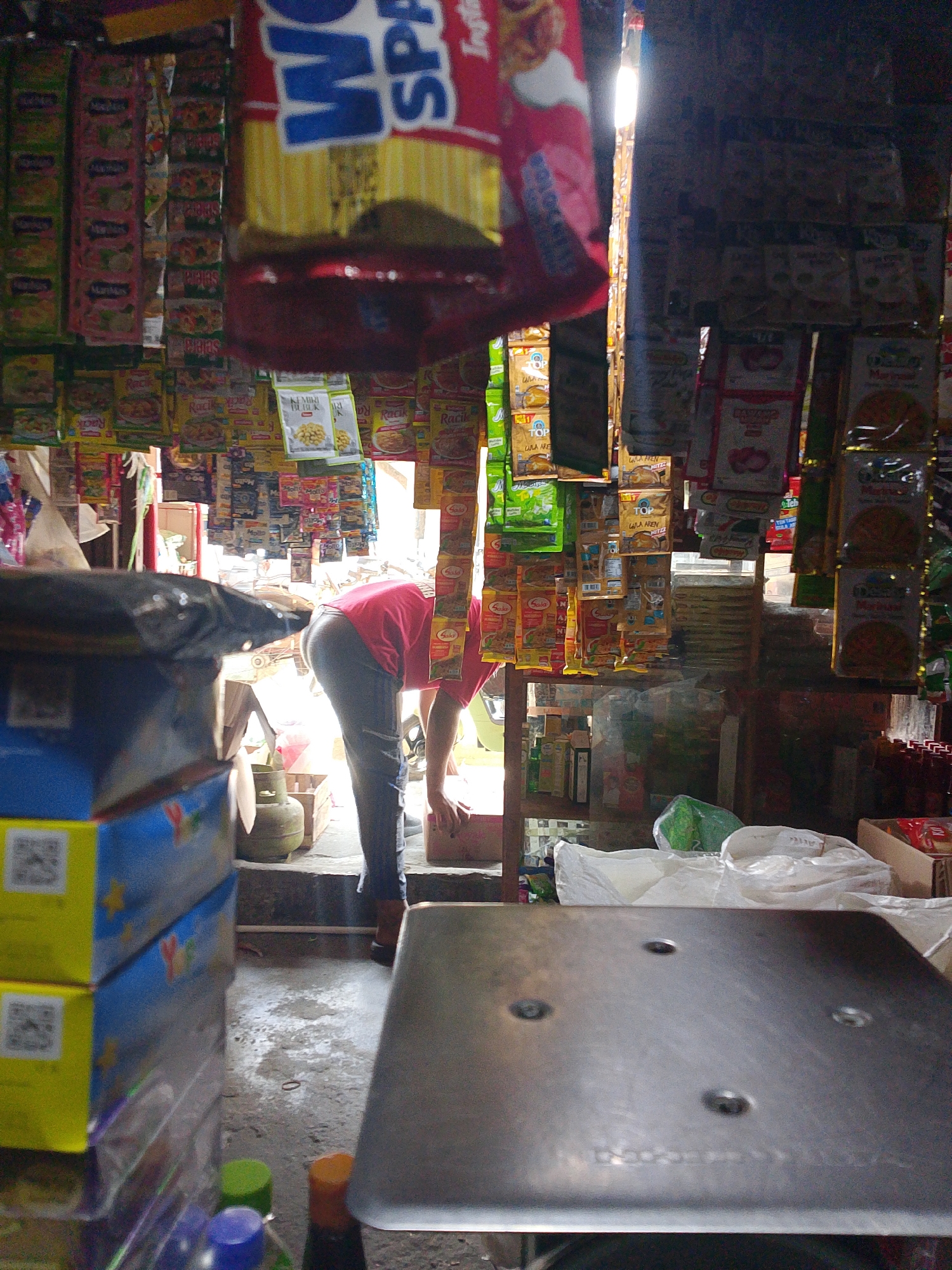 The image depicts a small, cluttered convenience store, likely a "warung," in Palembang, Indonesia. The interior is densely packed with various goods, primarily packaged food items and household necessities, hung from the ceiling and displayed on shelves. In the foreground, a stack of colorful boxes, possibly containing snacks or small toys, is visible. A person, wearing a red shirt and dark pants, is bent over in the middle of the store, attending to something on the floor or a low shelf. Their posture suggests they are in the process of stocking, retrieving, or arranging items. The lighting is uneven, with a bright light source, possibly sunlight or a strong bulb, illuminating the rear of the store and creating shadows. The floor appears to be concrete and somewhat damp or dirty. The overall impression is one of a busy, local retail establishment.