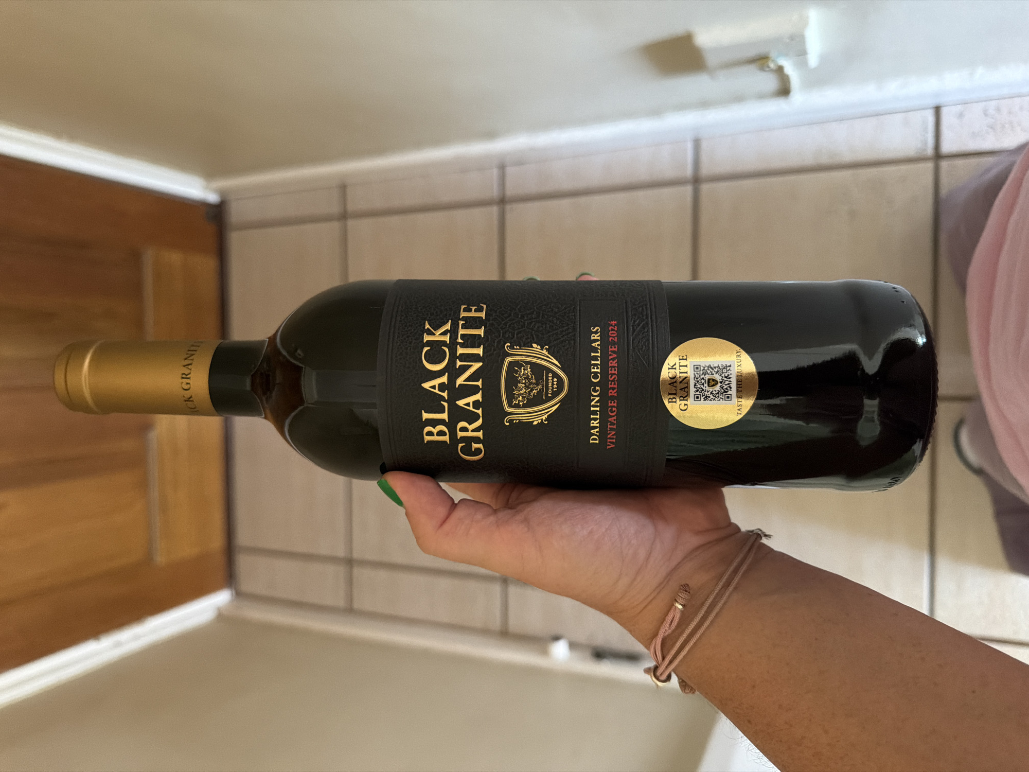 A person is holding a bottle of "Black Granite" red wine, a product of Darling Cellars, Vintage Reserve 2024. The bottle has a gold-colored cork and a black label with gold lettering. A gold sticker with a QR code is also visible on the bottle. The person's hand and a portion of their arm are visible, adorned with a pink bracelet. The background includes floor tiles, a portion of a light-colored wall, and a glimpse of a wooden door or furniture. Based on the label information referencing "Darling Cellars", the origin of the wine may be in the Darling wine region, which is about an hour outside of Cape Town, South Africa.