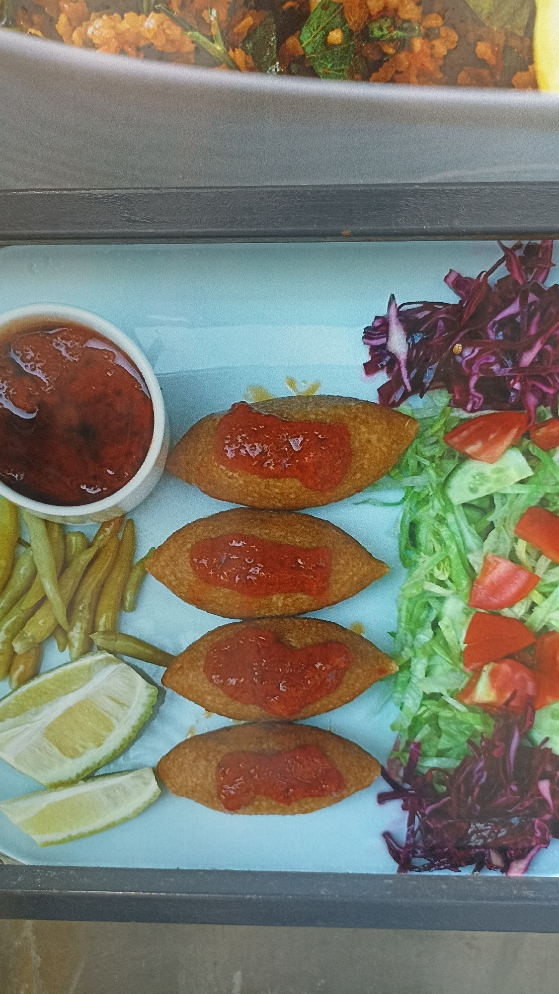 A plate of food is shown on a light blue surface. The plate contains four oval-shaped pieces of food, each topped with a dollop of red sauce. To the left of the food is a small white bowl filled with red sauce. Also on the plate are some green, thin, pickled vegetables and two slices of lemon. To the right of the food is a salad composed of shredded lettuce, chopped tomatoes, cucumber slices, and shredded red cabbage. The image is a close-up shot, and no people or specific city landmarks are visible.