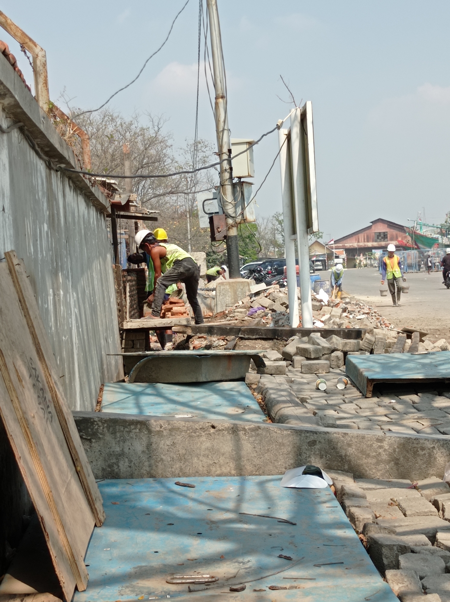 The image depicts a construction site in Tegal, Indonesia, characterized by debris and active work. In the foreground, a person wearing a yellow vest and a white hard hat is bent over, appearing to be engaged in some form of manual labor, possibly breaking or cutting materials. Another individual in a yellow hard hat and a lime green tank top is also working nearby. Behind them, a pile of bricks and construction materials is visible. Further in the background, two individuals in yellow vests are seen. One is sweeping, and the other is carrying two buckets. The environment includes a road with parked vehicles and motorcycles, as well as buildings in the distance. The overall scene suggests an active construction or demolition project in an urban setting.