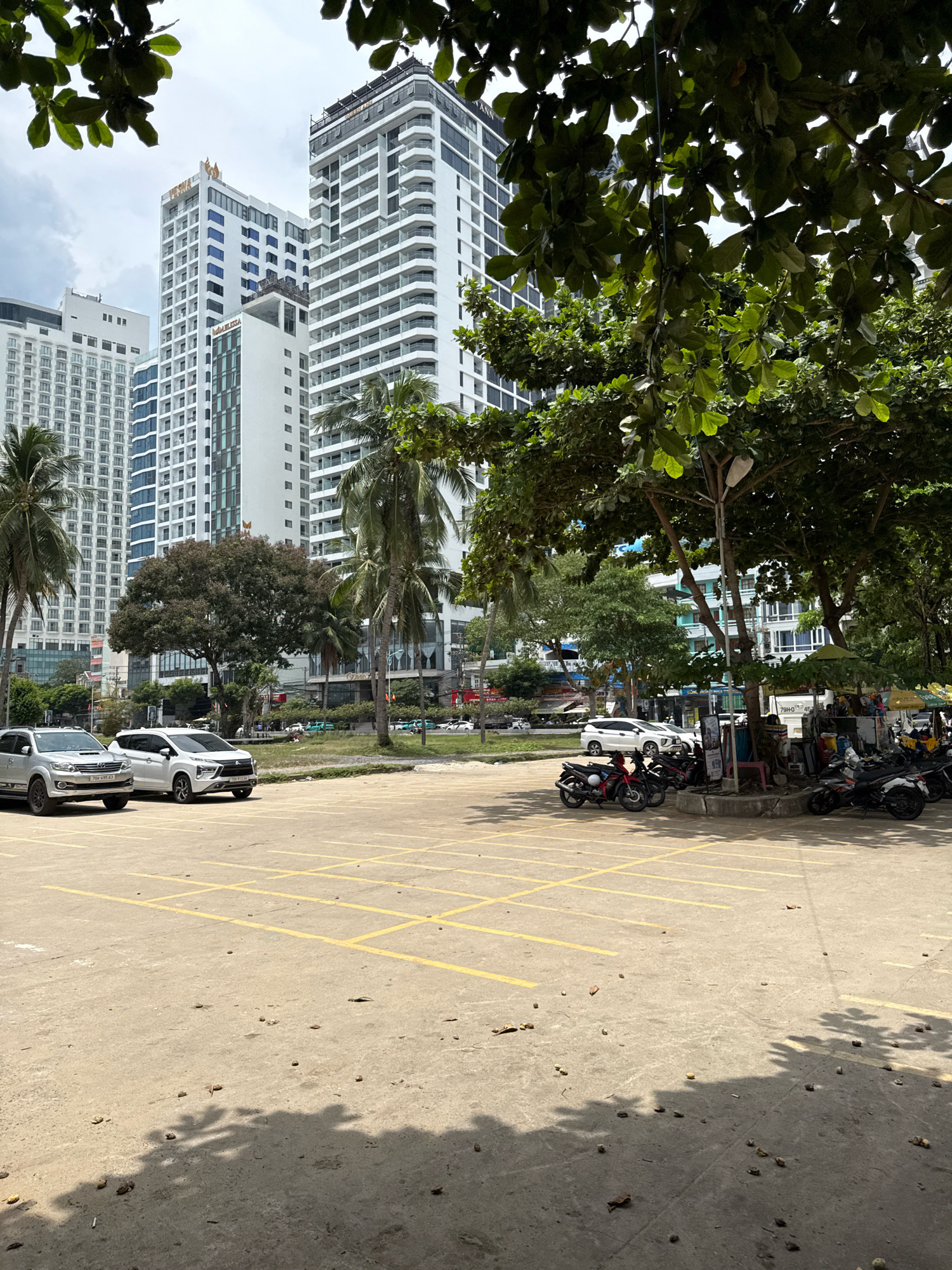 The image depicts a parking lot in Nha Trang, Vietnam, with several buildings in the background. The foreground features a concrete lot with yellow parking lines. Two cars, a silver SUV and a white SUV, are parked on the left side of the lot. A row of motorcycles is parked on the right side, some with riders. Beyond the parking lot, there is a street with more vehicles and a few people walking. Several tall buildings, likely hotels or apartment complexes, are visible in the background, characteristic of Nha Trang's coastal cityscape. Palm trees and other foliage are scattered throughout the scene, suggesting a tropical climate. The sky is overcast, with some patches of blue. The overall impression is that of a busy urban area on a cloudy day.
