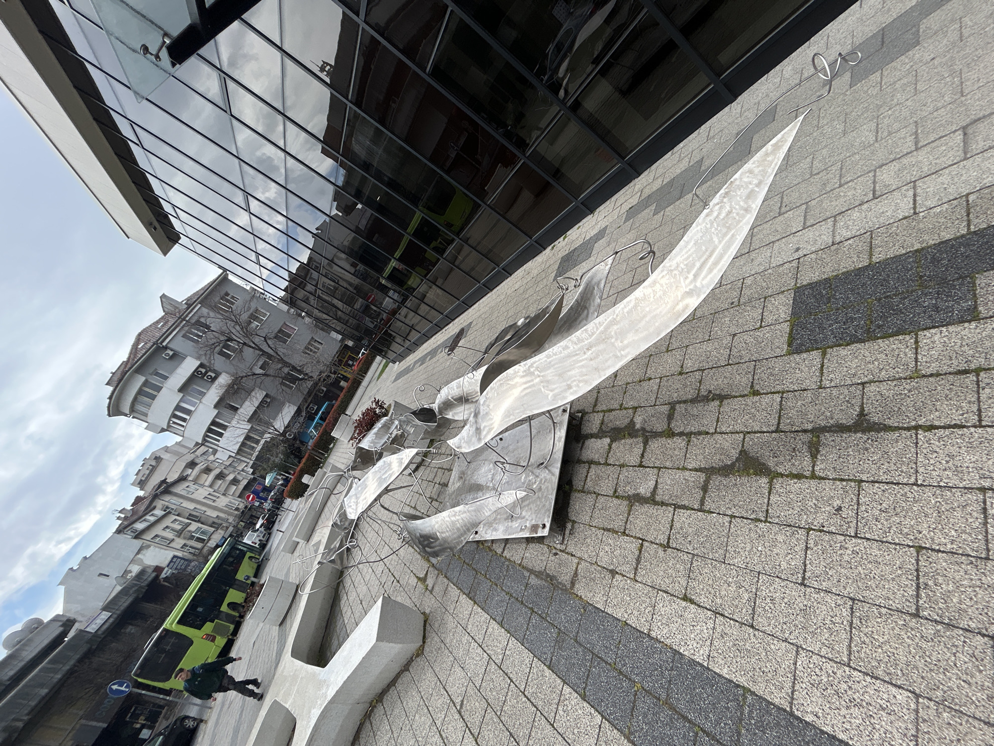 This urban scene in Plovdiv, Bulgaria, depicts a public square or pedestrian zone adjacent to a city street under an overcast sky. In the foreground, an abstract sculpture composed of multiple reflective metal sheets and coiled metal rods is installed on a square base plate, resting on light grey granite pavers. A linear strip of darker grey pavers runs diagonally across the lower left quadrant of the frame. Adjacent to the sculpture on the left, a large, light-colored concrete block, serving as seating or a planter base, is partially visible.
In the mid-ground, a lone male individual, dressed in a dark green jacket and dark trousers, walks away from the camera towards the upper left. Immediately behind this person, a bright green city bus with "177" displayed on its route sign is visible on a paved street. Beyond the street, a mix of older, multi-story residential and commercial buildings with varied facades are present, including structures with balconies and externally mounted air conditioning units. A blue circular traffic sign indicating a mandatory left turn and a red circular "no entry" sign are visible further down the street.
The right side of the frame is dominated by a modern building featuring a dark, highly reflective glass facade, which mirrors the cloudy sky and surrounding architecture. The overall environment is urban, characterized by architectural diversity and public infrastructure, under natural, diffused daylight.
