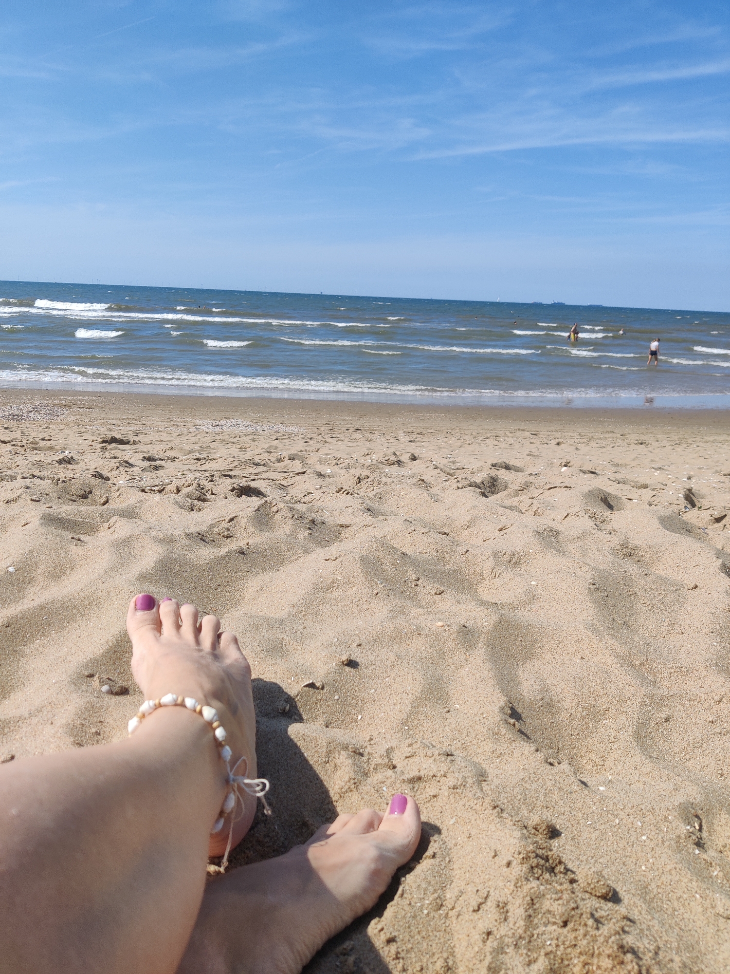 The image depicts a sandy beach under a clear blue sky with scattered clouds. The foreground shows a person's legs and feet resting on the sand. The feet are adorned with purple nail polish and a shell anklet. In the mid-ground, waves gently lap against the shore of the North Sea. Further out, several people can be seen wading or swimming in the water. The horizon line is visible, separating the sea from the sky, with faint indications of distant structures or ships. This scene is characteristic of the beach at Scheveningen, a popular seaside resort in The Hague, Netherlands, known for its vast sandy beaches and recreational activities.