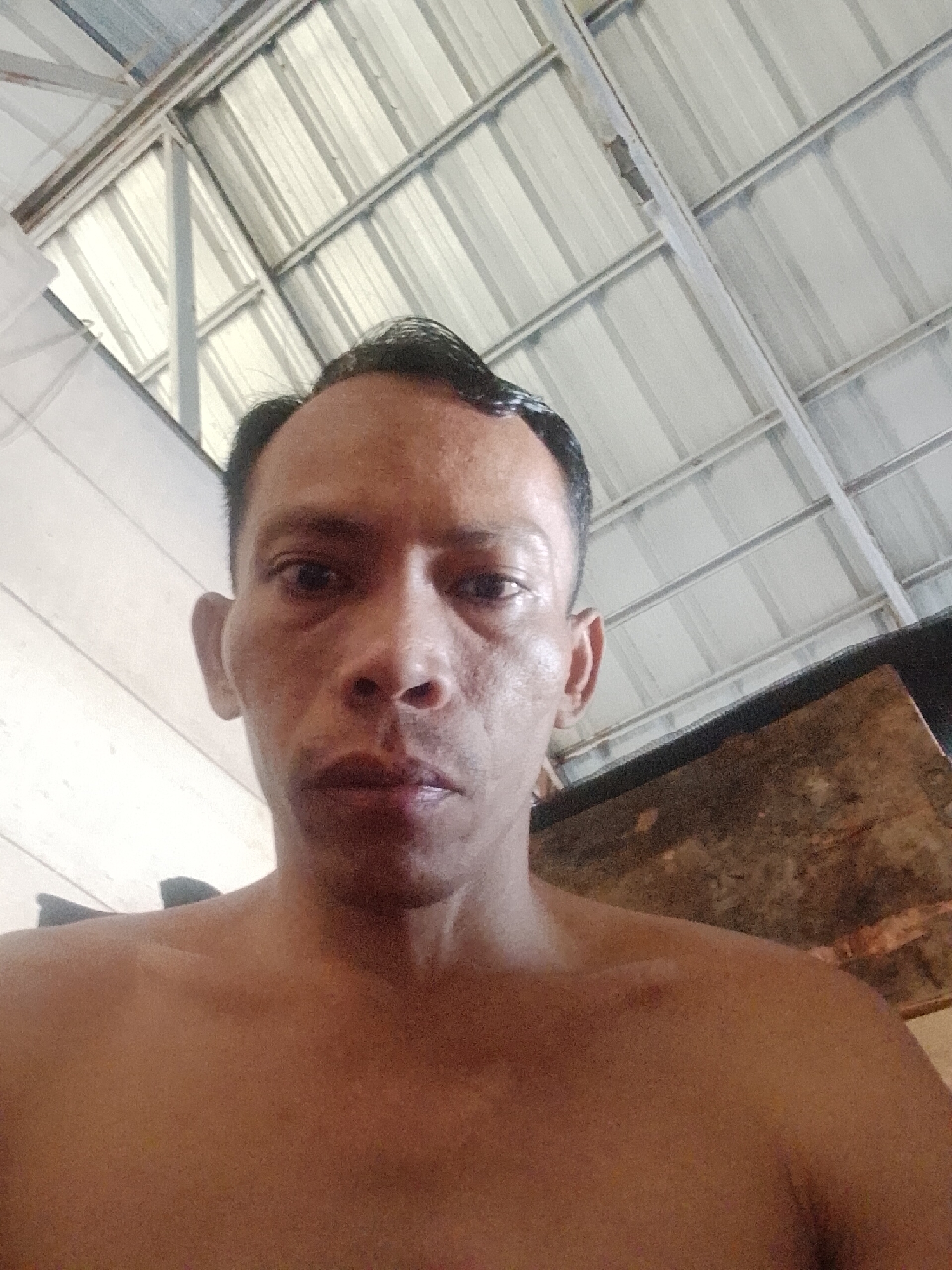A close-up, low-angle, unflattering selfie of a shirtless middle-aged man with dark, slicked-back hair. He is fair-skinned with visible pores and wrinkles. The background features a corrugated metal roof with a supporting framework of white-painted beams. To the right, there is a dark, textured wooden surface. The image was taken in Bukit Ketok, Indonesia.
