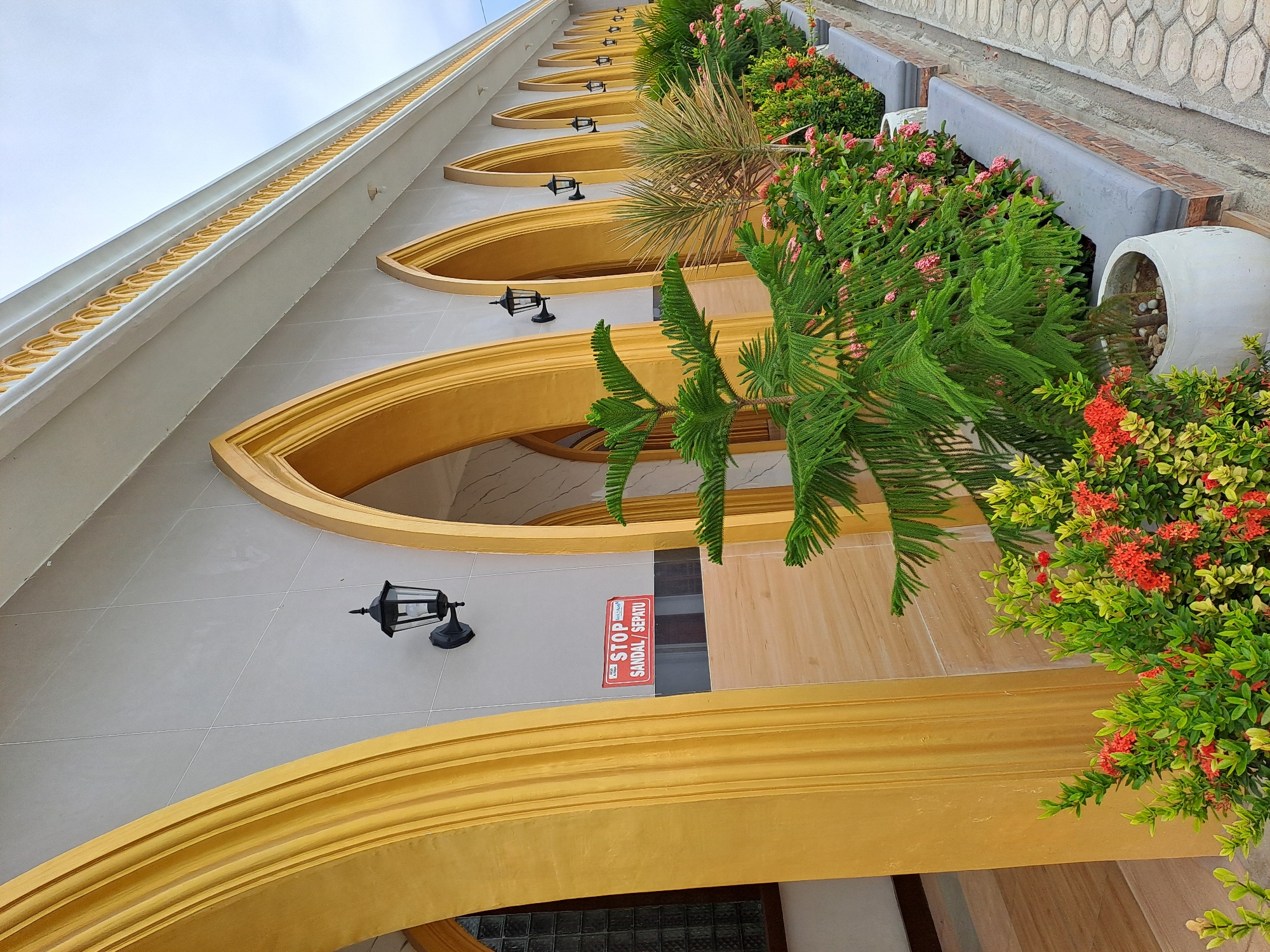 The image shows the exterior of a building in Lhokawe Teungoh, Indonesia. The building features a row of arched openings with gold trim, and a light grey tiled facade. Black outdoor lamps are mounted on the wall between each arch. A red and white "STOP SANDAL/SEPATU" (Stop Sandals/Shoes) sign is visible on the wall to the left. In front of the building, a variety of potted plants and flowers are arranged along a raised planter. These include a large evergreen tree, a spiky palm-like plant, and flowering bushes with red and pink blossoms. A paved walkway with a hexagonal pattern runs alongside the planter. The sky is overcast.