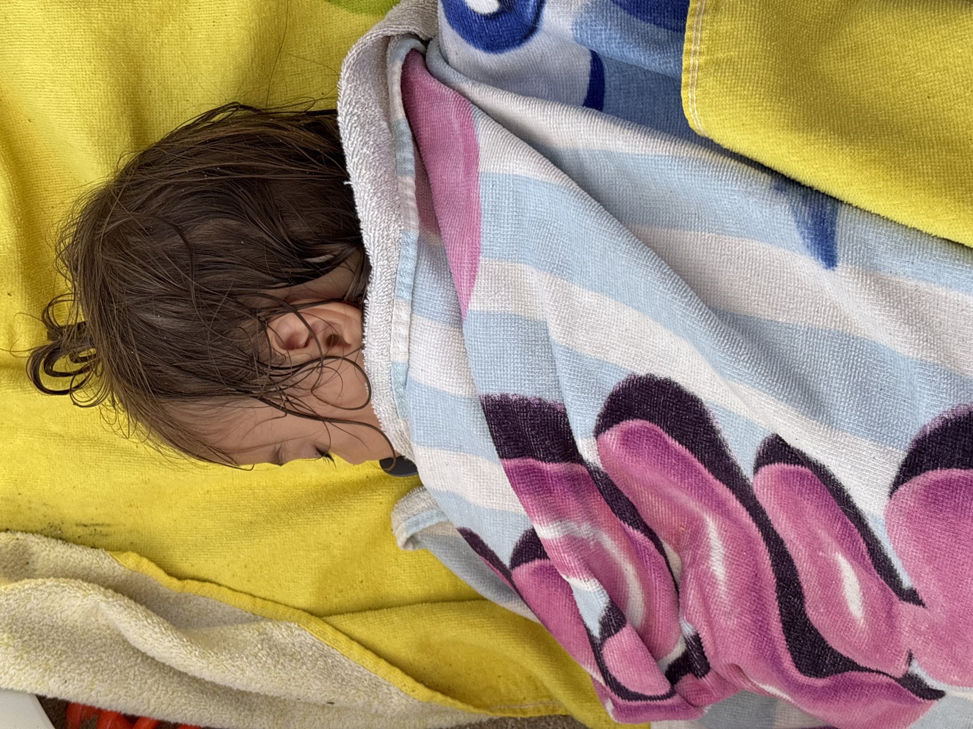 A close-up, overhead shot shows a baby sleeping on a yellow towel, wrapped in a colorful towel with stripes and a floral pattern of pink and purple. The baby's head is turned to the side, with wet, dark hair styled in a messy bun. The baby is wearing a pacifier. The scene appears to be outdoors, possibly at a beach or pool area, given the towels and the bright yellow background, which could be a beach towel. The image is taken in Primorsko, Bulgaria, a seaside resort town known for its beaches.