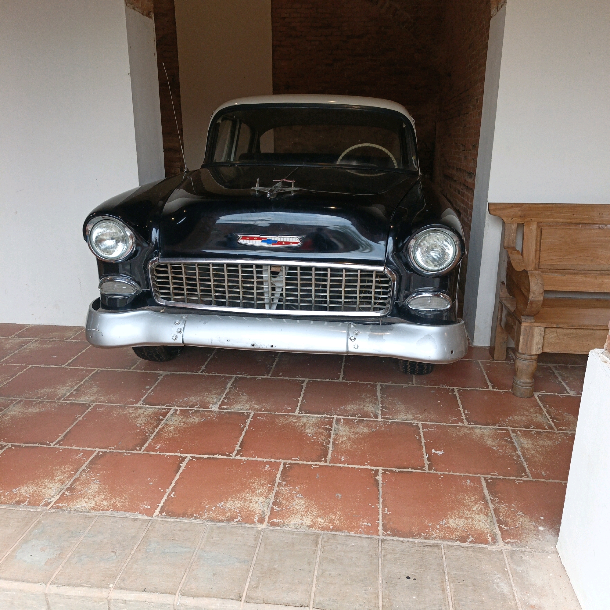 An antique black Chevrolet Bel Air, a classic car, is parked indoors, likely in a covered area. The car has a white roof and a chrome grille. The floor is tiled with red and grey squares, and a wooden bench is visible to the right. The background consists of brick walls and white pillars. The scene is devoid of people. This image was taken in Bejalen, Indonesia.