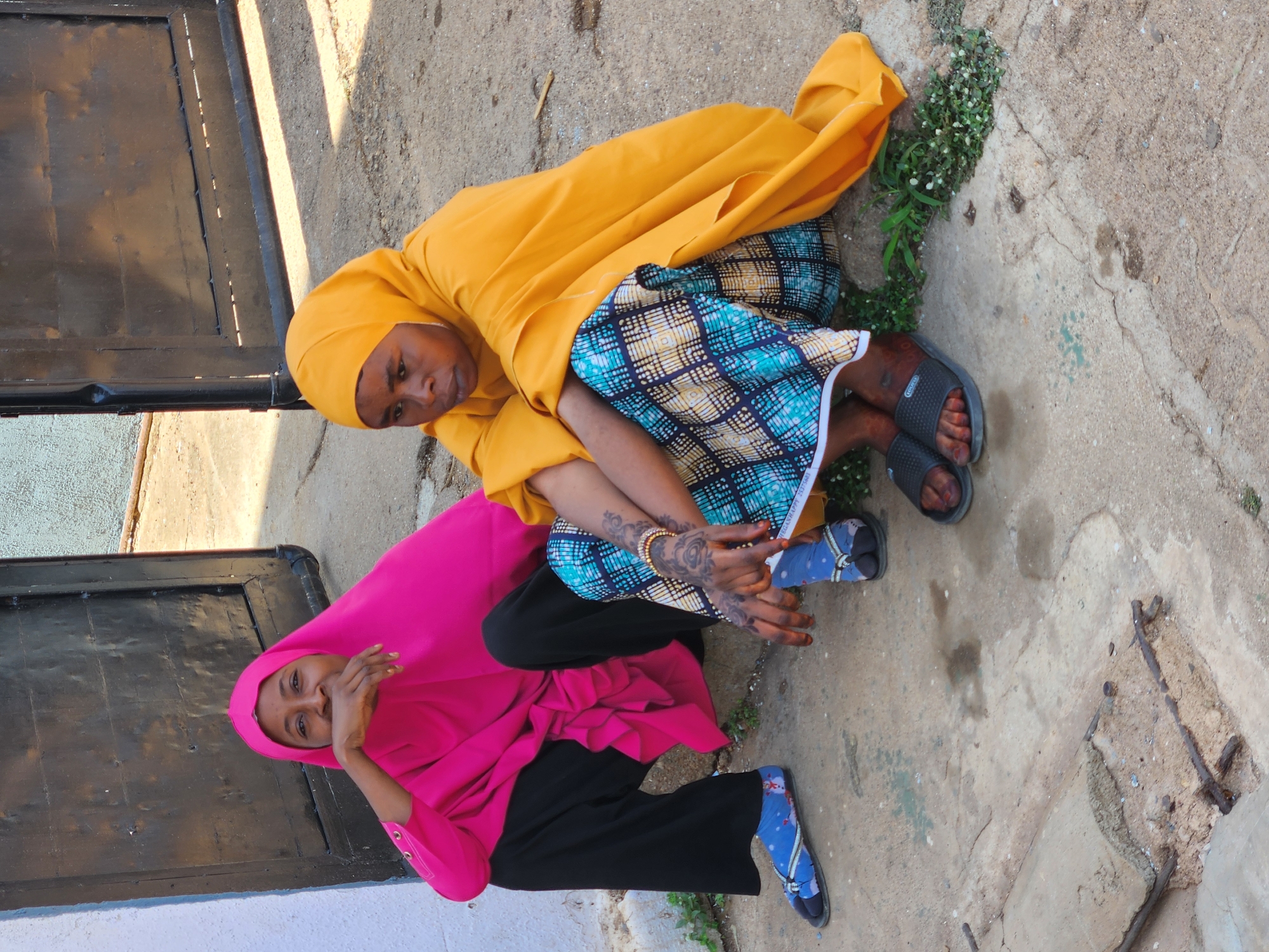 Two women are seated on the ground outdoors in Unknown, Unknown. One woman wears a yellow headscarf and matching top, along with a blue and yellow patterned skirt. She has tattoos on her arms and wears black sandals. The other woman wears a pink headscarf and matching top, with black pants and blue patterned socks with sandals. Both are near a dark-colored door on a concrete surface. There is some greenery visible around them.