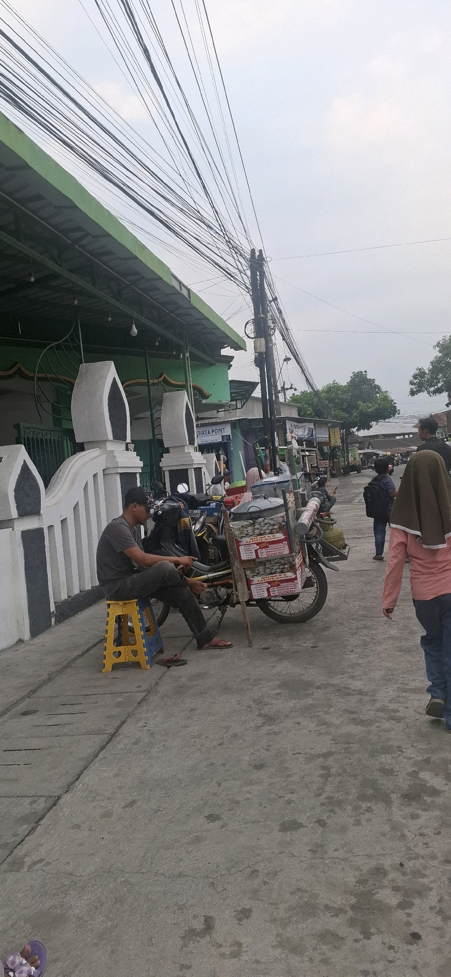 In Desa Dibal, Indonesia, a street scene unfolds under an overcast sky. A man wearing a dark t-shirt, dark pants, and a black cap sits on a yellow and blue folding stool beside a motorcycle. The motorcycle is equipped with a cart carrying several stacked boxes, each labeled with text including "JAMU" and "JAMUR." A single gas cylinder is also visible in the cart.
To the left of the man, a low white wall with black accents runs along the sidewalk, leading to a building with a green roof and green walls. Decorative white pillars with curved tops are visible along the wall. Overhead, a dense network of black electrical wires crisscrosses the sky, supported by a tall utility pole.
Further down the street, a woman wearing a pink long-sleeved shirt and blue jeans walks away from the camera, her head covered by a brown hijab. Another person in a dark backpack is seen walking in the distance. Several other motorcycles and a green truck are also present on the street. The ground is paved with concrete, showing signs of wear and some scattered dark stains. The overall atmosphere is one of everyday street life in a local Indonesian town.