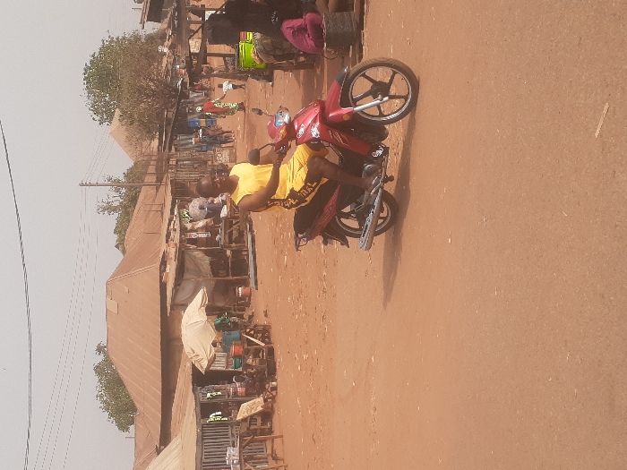 The scene depicts an unpaved, reddish-brown dirt road in Gwadanye, Nigeria, under a hazy, light-colored sky. A male individual, dressed in a yellow sleeveless shirt and yellow shorts, is seated centrally on a stationary red motorcycle. He is looking towards the viewer with a slight smile; his bare feet rest on the motorcycle's footpegs. The motorcycle features black wheels and a chrome exhaust pipe on the right side.
In the background, a bustling marketplace unfolds with numerous makeshift stalls constructed from wooden frames and various roofing materials, including corrugated iron sheets and fabric awnings. Several people are visible among these stalls, some sitting or standing. One individual in a distinctive patterned dress is walking towards the right, and a child is near them. Larger, more permanent structures with reddish-brown corrugated metal roofs are situated behind the stalls. Sparse trees with light foliage are scattered throughout the background. Overhead, multiple power lines traverse the scene. The overall environment appears dry and dusty.