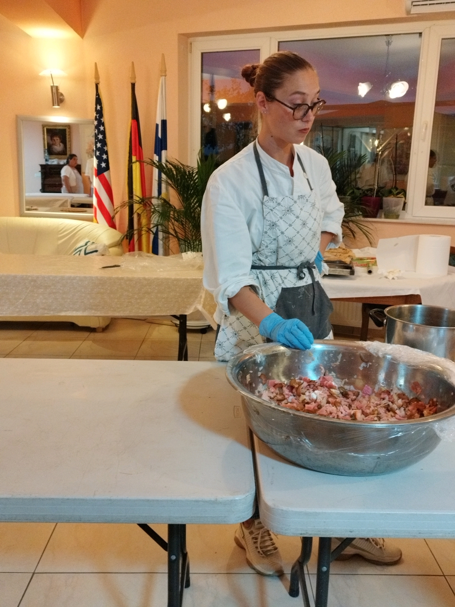 In Bucharest, Romania, a woman wearing glasses, a white shirt, and an apron with blue gloves is preparing food in a stainless steel bowl. The bowl contains a mixture of chopped ingredients, possibly meat and vegetables, and is covered with plastic wrap. To the right of the bowl is a large metal pot. The woman is standing behind a white folding table, and another folding table is visible in the foreground. In the background, several flags are displayed, including the American flag, the German flag, and the Finnish flag. A sofa and a framed portrait are visible in the reflection of a mirror on the left side of the image. Plants are also present in the background, and a window shows a sunset. The room has orange-colored walls and a tiled floor.