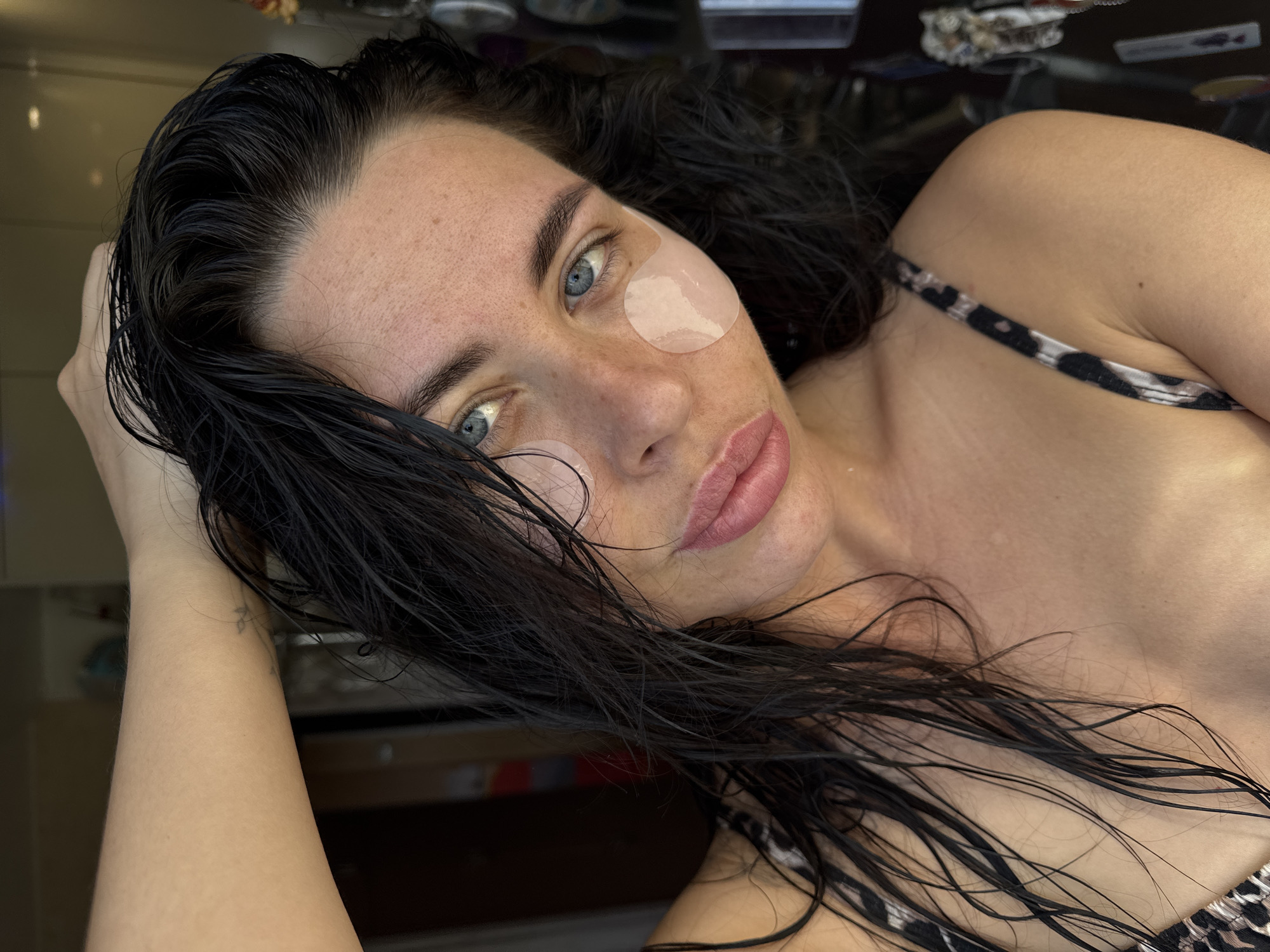 The media file presents a close-up portrait of a woman, seen from an elevated angle, lying down and looking directly into the camera. She has long, dark hair that appears wet, fanned around her head and shoulders. Her eyes are a striking blue, and her fair skin shows a scattering of freckles, particularly on her forehead and cheeks. Two light-colored, translucent patches, likely under-eye masks, are placed beneath each of her eyes, suggesting a skincare or relaxation activity. Her full lips are slightly parted, revealing a natural pink hue. She is wearing a top with a black and white leopard print strap, visible on her right shoulder. Her right arm is bent, with her hand supporting her head, and a faint tattoo is visible on her left wrist. The background is dark and out of focus, with some reflective surfaces indicating an indoor environment, possibly a bathroom or bedroom. The woman's expression is calm and direct. There are no specific cues for time of day or weather, and no visible text is present. This scene is set in Chaikovsky, Russia.