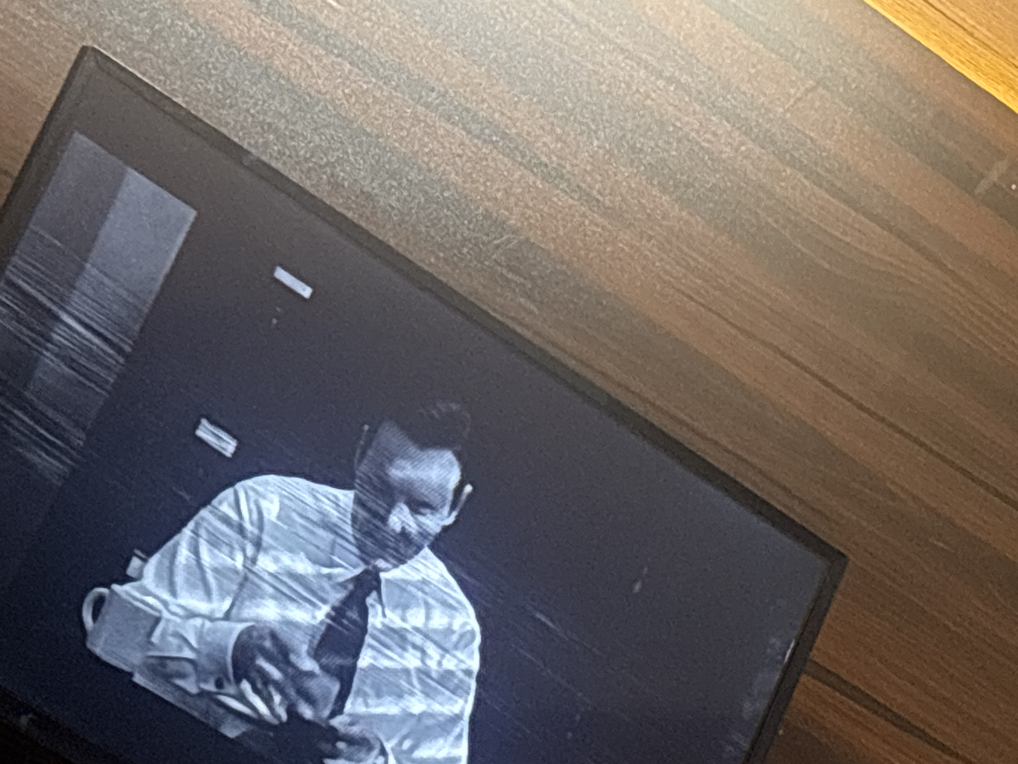 The image shows a television screen displaying a black and white film or video. A man in a light-colored, patterned shirt and tie is visible on the screen, sitting at what appears to be a desk. The screen has vertical lines or distortion. The television is placed on a wooden surface. The location is not discernable from the visual information.