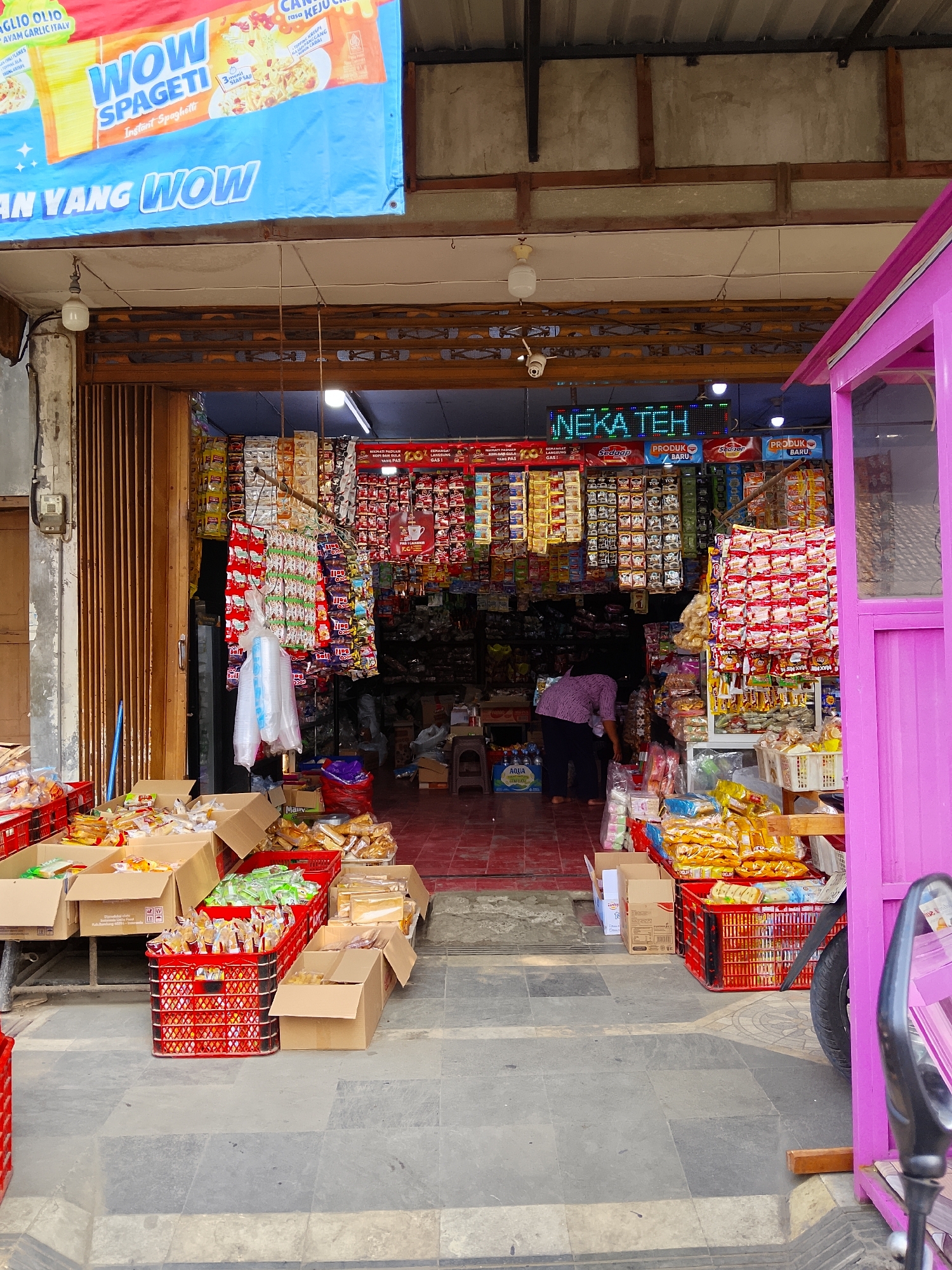 The image depicts the exterior of a small convenience store, identified as "NGEKA TEH", located in Ngelo, Indonesia. The store appears to be open for business, with its entrance wide open, revealing shelves stocked with various food items and packaged goods. A large blue banner with text is visible above the entrance, featuring images of spaghetti. Red plastic crates filled with products are stacked on the ground in front of the store, along with cardboard boxes. A person, wearing a purple top and dark pants, is visible inside the store, seemingly attending to the inventory. To the right, a bright pink structure, possibly a kiosk or an extension of the store, is prominent. The ground is paved with grey tiles. The overall impression is of a bustling local shop in a residential or commercial area.