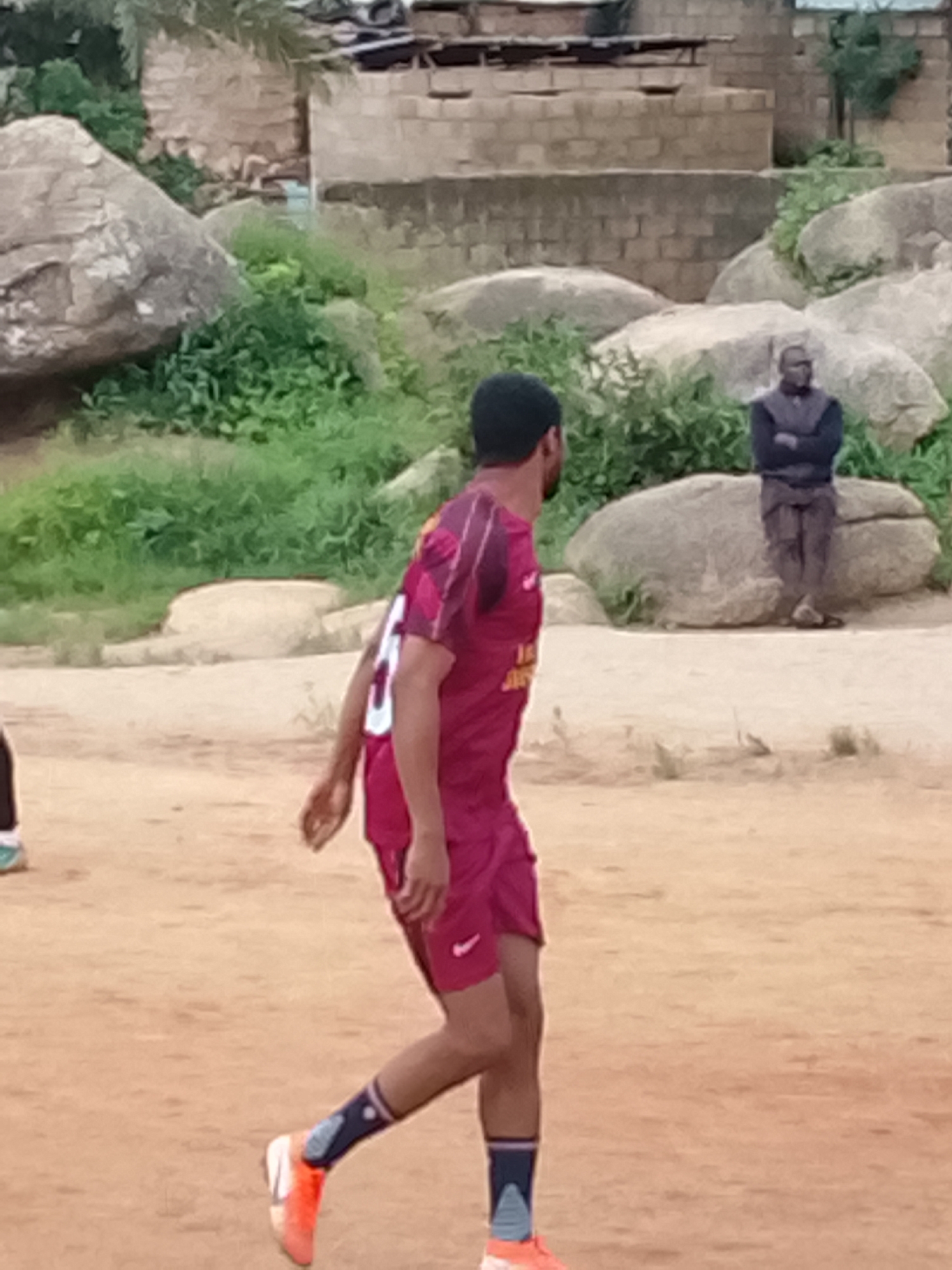In Jos, Nigeria, two men are visible in a dusty, outdoor setting. One man, wearing a maroon jersey with white lettering and numbers, and maroon shorts, stands in the foreground, facing away from the camera. He has dark hair and is wearing orange athletic shoes with dark blue and light blue socks. The other man, in the background, is seated on a large rock. He is wearing a dark vest over a light-colored shirt and dark trousers, with his arms crossed. The environment includes large, grey rocks, green vegetation, and some rudimentary brick structures in the distance, suggesting a rural or semi-urban area. The ground appears to be dry earth or sand.