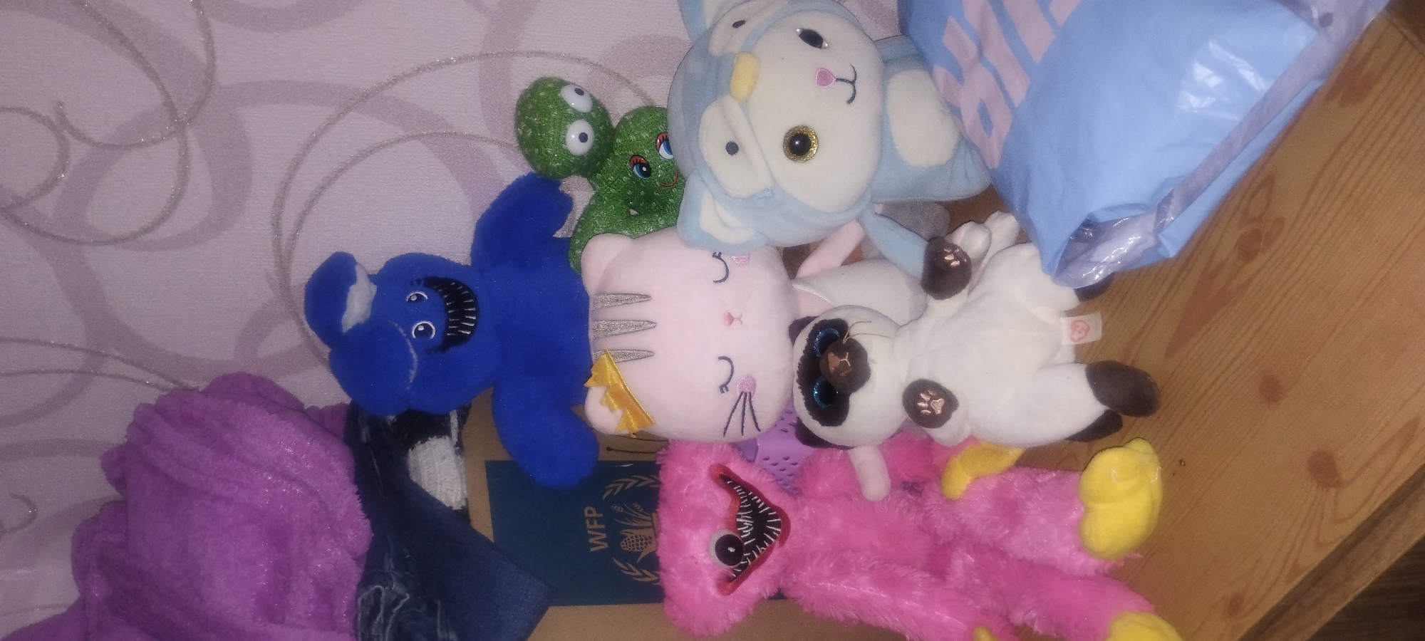 The image displays a collection of stuffed toys arranged on a wooden surface, likely a shelf or dresser. In the background, a wall with a textured wallpaper pattern is visible. A portion of what appears to be a document with the "WFP" logo is also present. The stuffed toys include a large pink fuzzy monster with sharp teeth, a blue character with a similar toothy grin, a light pink cat with a yellow crown, a Siamese cat plush, a small green creature with googly eyes, and a light blue animal-themed plush. A folded light blue plastic bag is situated in the foreground. The lighting suggests an indoor environment. No individuals are present in the image. There is no information in the image that could indicate the location as Zaporizhzhia, Ukraine.