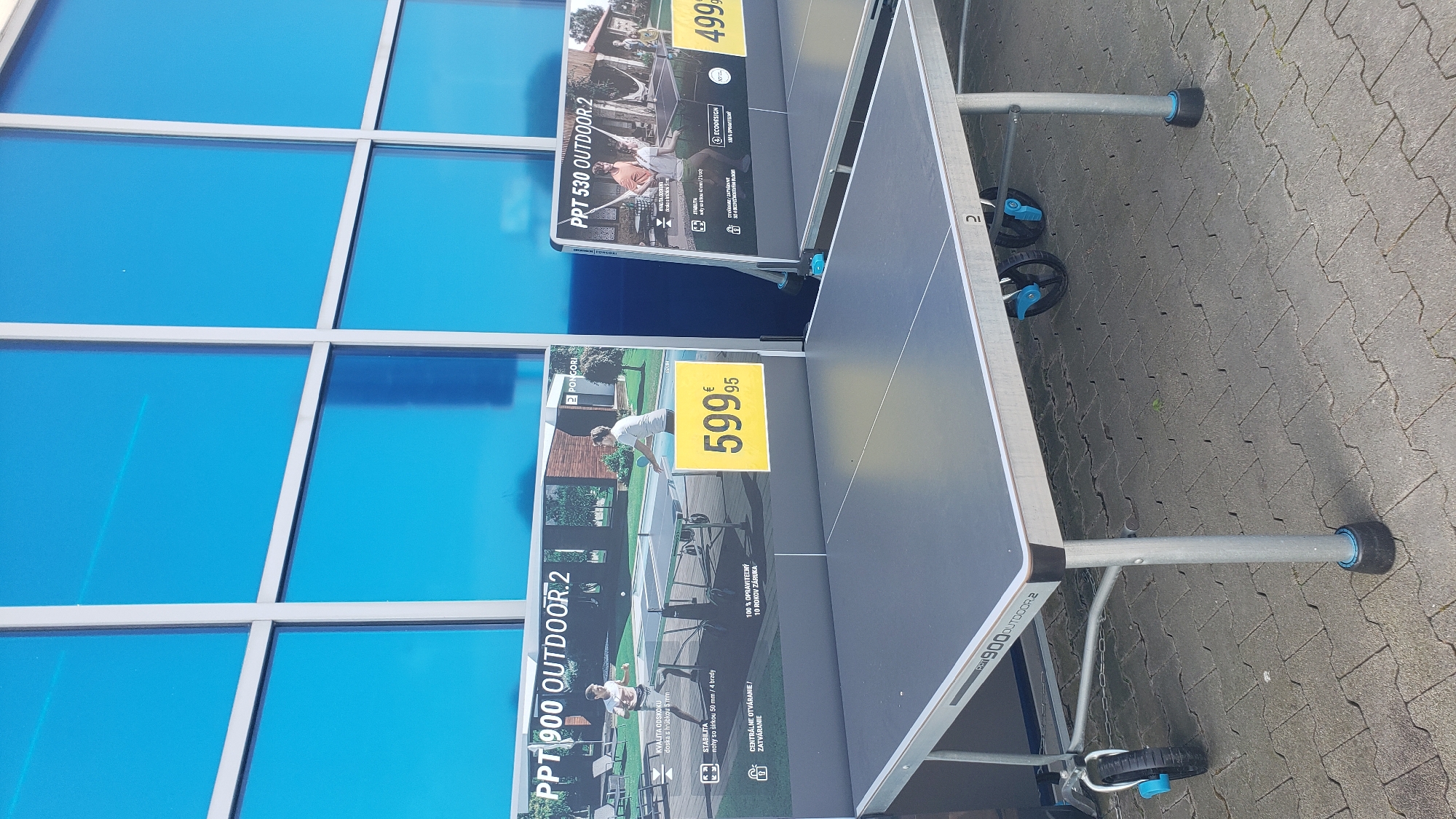 The image shows two outdoor table tennis tables displayed in front of a building with a blue, grid-patterned glass facade. The building appears to be a retail establishment, possibly a sports store, located in Prešov, Slovakia. The table in the foreground is a gray "PPT 900 OUTDOOR.2" model. To the left of this table, a sign displays an advertisement for the "PPT 900 OUTDOOR.2" table tennis table, featuring an image of two people playing and the price "599,95 €". To the right, partially visible, is another table tennis table, likely the "PPT 530 OUTDOOR.2", also with a sign indicating its price is "499 €". Both tables have metal legs and wheels, suggesting they are portable. The ground is paved with interlocking gray stones. No people are actively interacting with the tables. The overall environment is an outdoor display area of a commercial building.