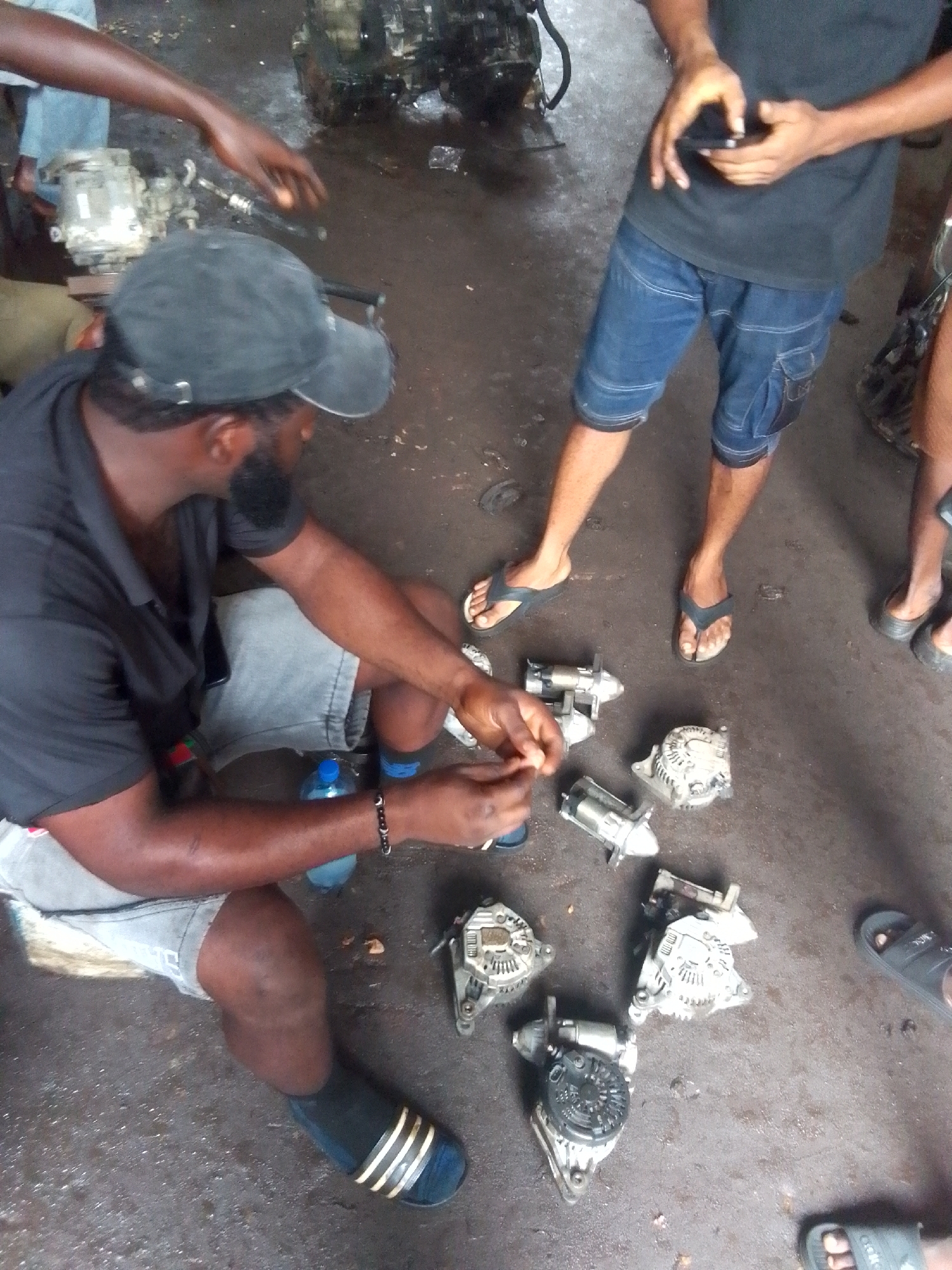 The scene depicts an automotive repair shop in Onitsha, Nigeria. There are four individuals present. One man, wearing a black shirt, grey shorts, and a black cap, sits on the floor examining several car alternators. Another man stands nearby, wearing a black shirt and blue jean shorts, looking at his phone. Two other individuals are partially visible. The floor is dirty and has auto parts scattered around. In the background, there's an engine block and various automotive components. The setting suggests a small, informal auto repair business environment.