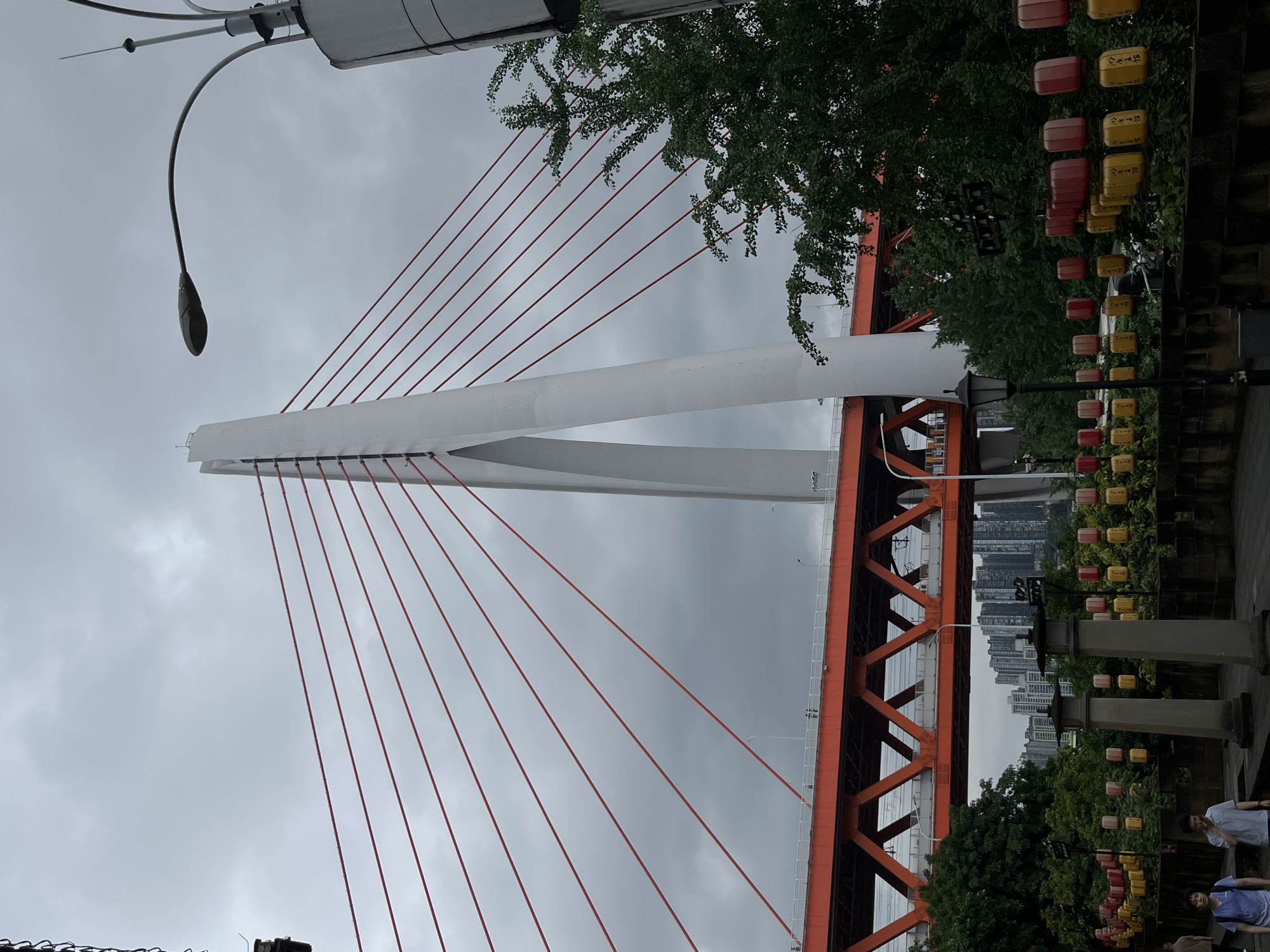 The image depicts the Chongqing Bridge, a cable-stayed bridge located in the Yuzhong District of Chongqing, China. The bridge is characterized by its distinctive white pylon with a wishbone shape. Red cables extend from the pylon to support the orange bridge deck. The sky is overcast and gray. The bridge spans a river, with a cityscape visible in the background. In the foreground, there is vegetation, lanterns, and a few people. The lanterns are red and yellow. A street light is also visible.