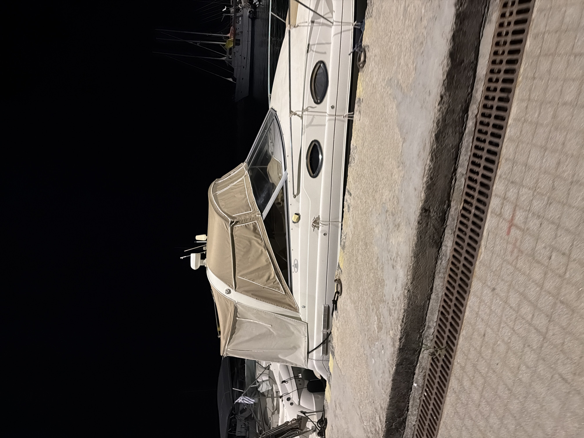 A white yacht is docked at night in the Municipal Unit of Alyzia, Greece, at a pier with a concrete surface and a metal grate drain. The yacht has a tan canopy extending over the cockpit area. A smaller boat with a black canopy is docked behind it, featuring a Greek flag. In the background, other boats and masts are visible in the dark water. The scene appears still and quiet, illuminated by artificial light.