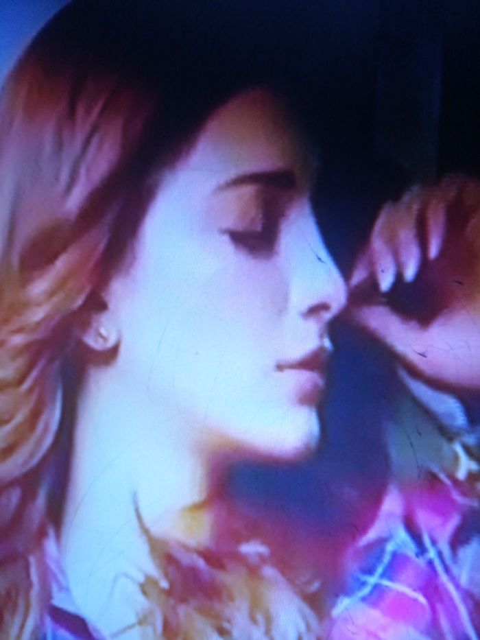 A woman's profile dominates the frame. She has fair skin, dark hair, and closed eyes. Her nose is prominent and she has full lips. A hand partially obscures her face, fingers delicately touching her cheek. The background is dark, and the overall lighting is blue-tinged, casting shadows on her face. A plaid pattern is faintly visible on the lower right, suggesting clothing. The image has a soft, romantic quality.