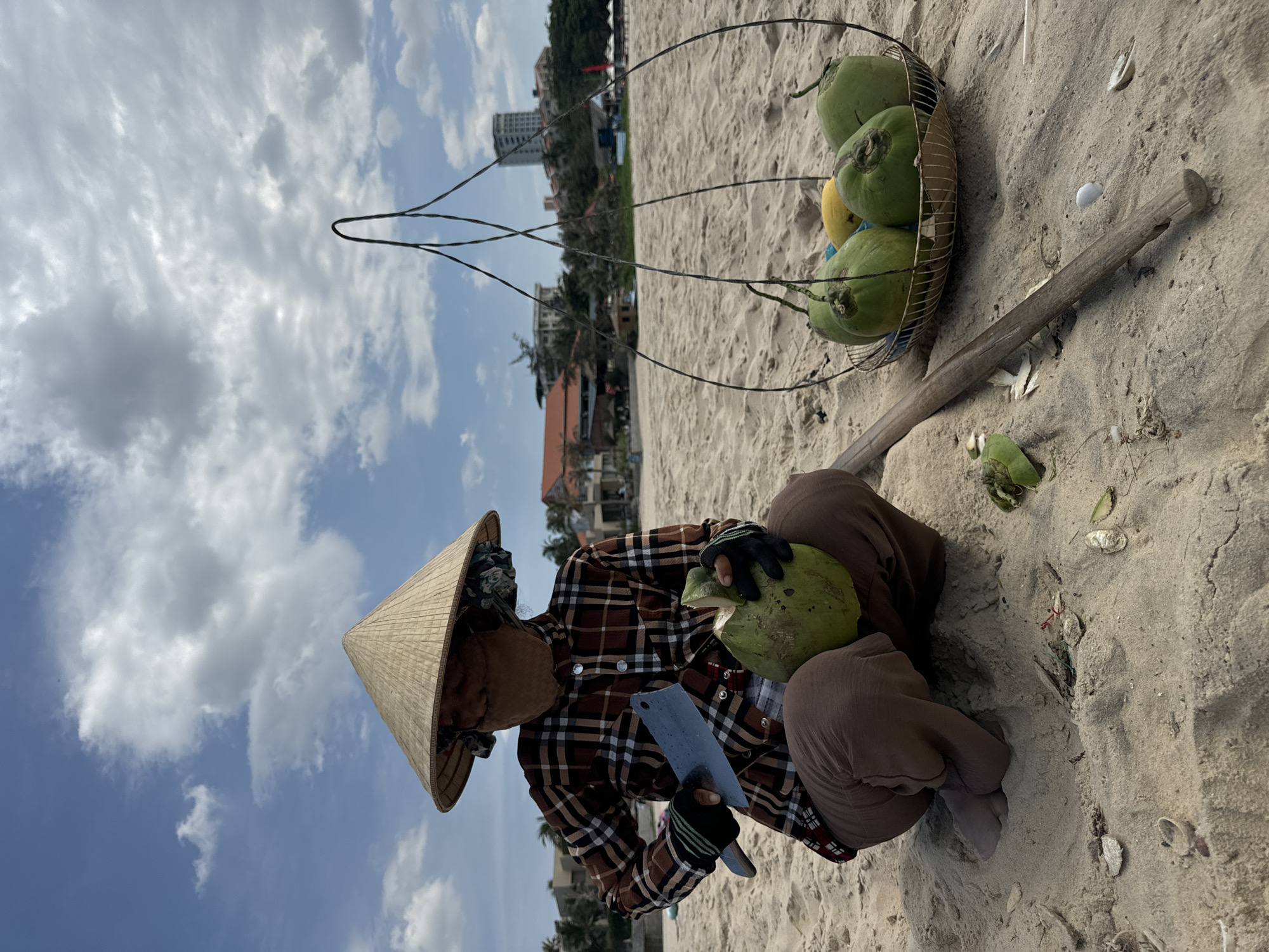A woman wearing a traditional conical Asian hat and a plaid shirt sits on a sandy beach. She is using a large knife to open a green coconut. Beside her is a wire basket containing several more coconuts. A wooden stick rests on the sand next to the basket. The location appears to be a beach, possibly in Phu Thuy Ward, Vietnam, based on the surroundings. The sky is partly cloudy. The woman is focused on opening the coconut, suggesting she may be a vendor selling them to tourists.