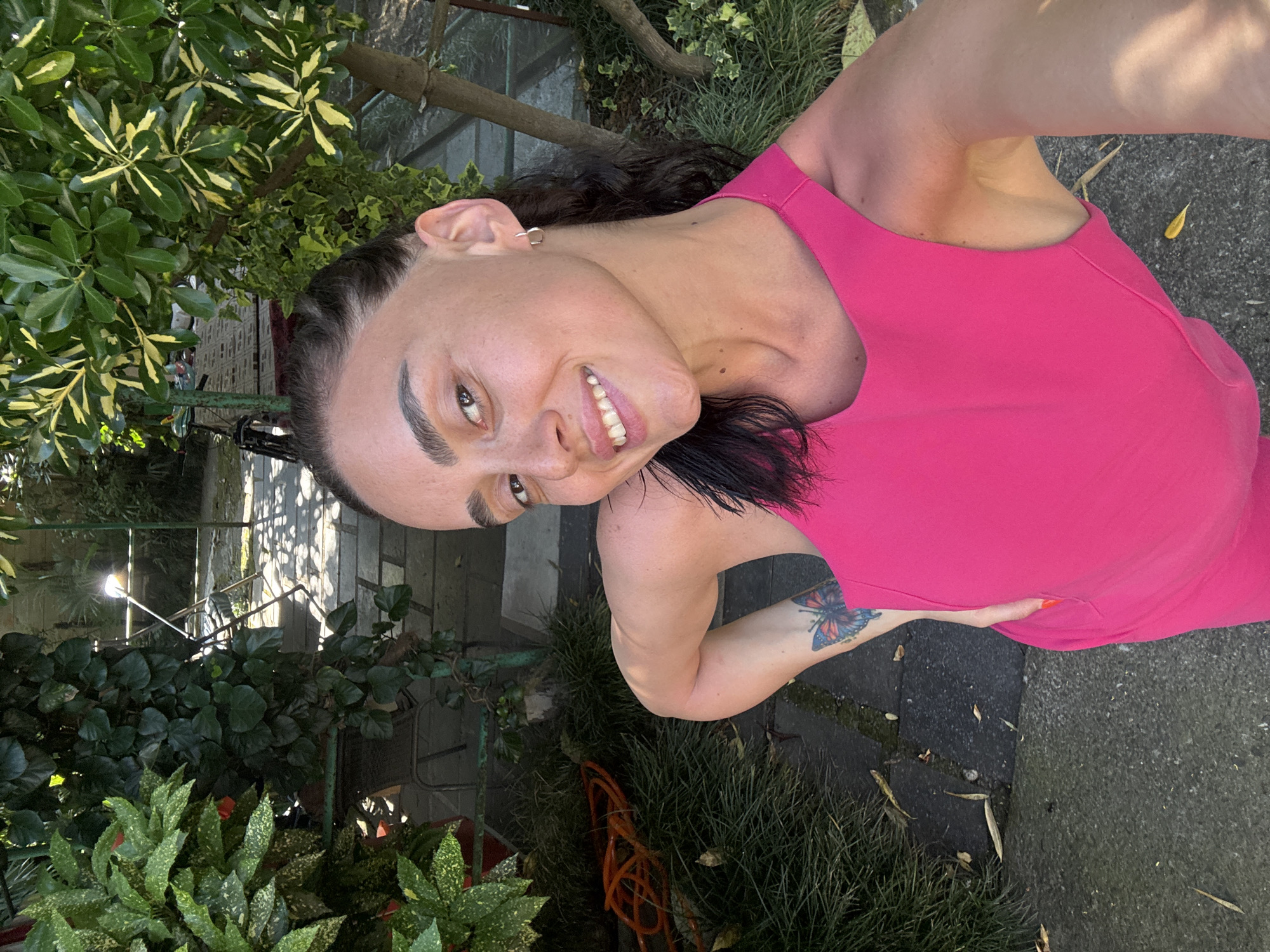 A woman with dark hair pulled back smiles at the camera, taking a selfie. She is wearing a bright pink sleeveless top. She has a butterfly tattoo on her left arm. She is standing outdoors in what appears to be a garden or courtyard. There are lush green plants and trees surrounding her. Sunlight is filtering through the leaves, creating dappled shadows on the ground. In the background, a table and chairs are visible, as well as a paved walkway. The overall impression is one of a sunny, relaxed day in a well-maintained outdoor space.