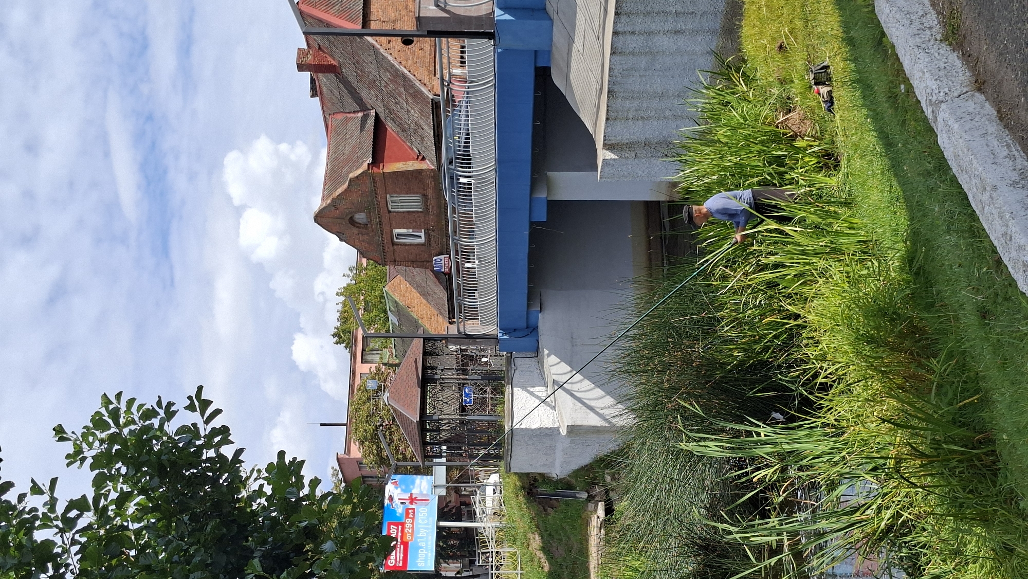 An elderly male is fishing with a rod in a narrow canal or stream in Pastavy, Belarus. He is standing amidst thick reeds and grass on the bank of the water. A blue bridge with white railings spans the canal in the background. Buildings with red tiled roofs and brick facades are visible behind the bridge, as well as a white billboard with an advertisement. The sky is partly cloudy, with white clouds against a blue background. The lighting suggests it is daytime, likely afternoon, with sunlight casting shadows. A fishing bag is placed on the grassy bank near the fisherman.