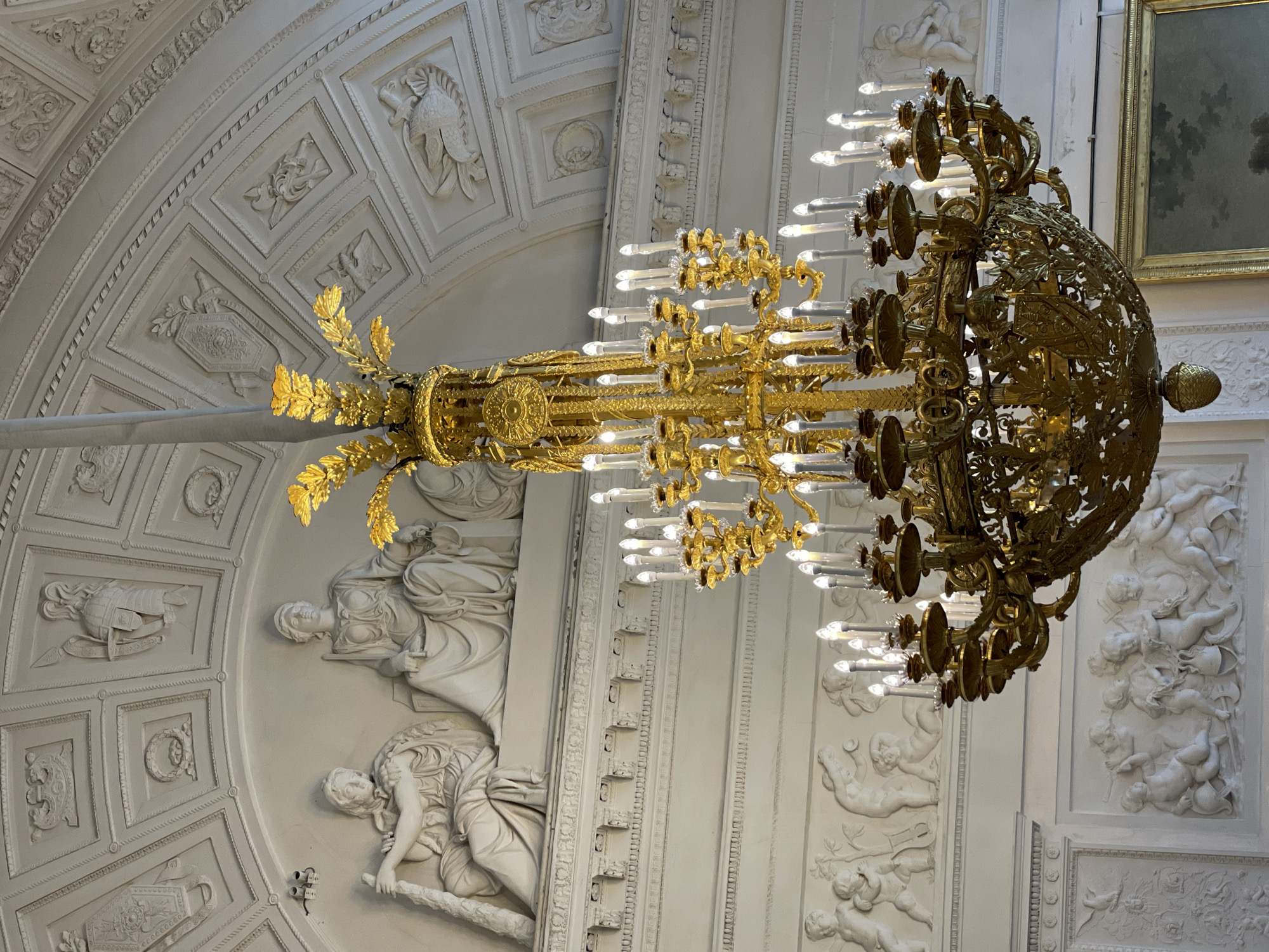 The photograph captures a detail of an interior, focusing on a large, ornate chandelier against a backdrop of classical architectural elements. The chandelier is predominantly golden in color, featuring multiple tiers and adorned with numerous light fixtures, some of which are illuminated. Its design incorporates intricate patterns and decorative elements, suggesting a high level of craftsmanship.
The background reveals a vaulted ceiling embellished with relief sculptures and moldings. The sculptural elements depict human figures in classical poses, surrounded by intricate decorative patterns and panels. The overall style of the architecture and the chandelier suggests a historical setting.
The image does not contain any immediately discernible information regarding the location or individuals present. The city remains unknown.