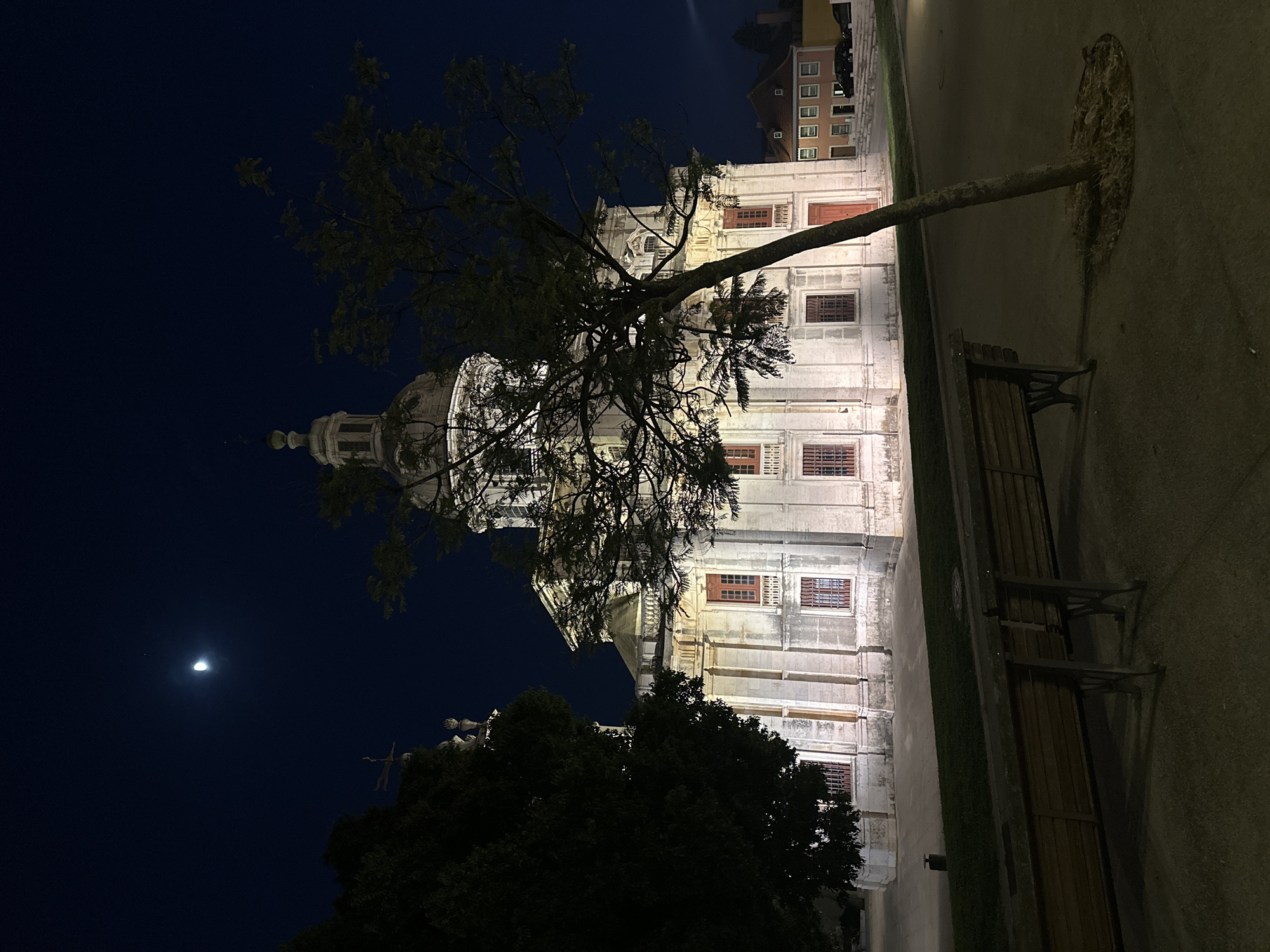 The photograph depicts a nighttime scene of the Mafra National Palace, located in Mafra, Portugal, just outside of Lisbon. The imposing white stone edifice, characterized by its grand dome and classical architecture, is illuminated by artificial lights, highlighting its ornate facade with rows of windows and a prominent red door. A half-moon is visible in the dark, clear night sky. In the foreground, a dark tree with bare branches leans across the frame, partially obscuring the palace. A wooden bench with metal legs sits on a paved walkway, in front of a strip of grass. Further in the background, several other buildings with visible windows and a car can be observed.