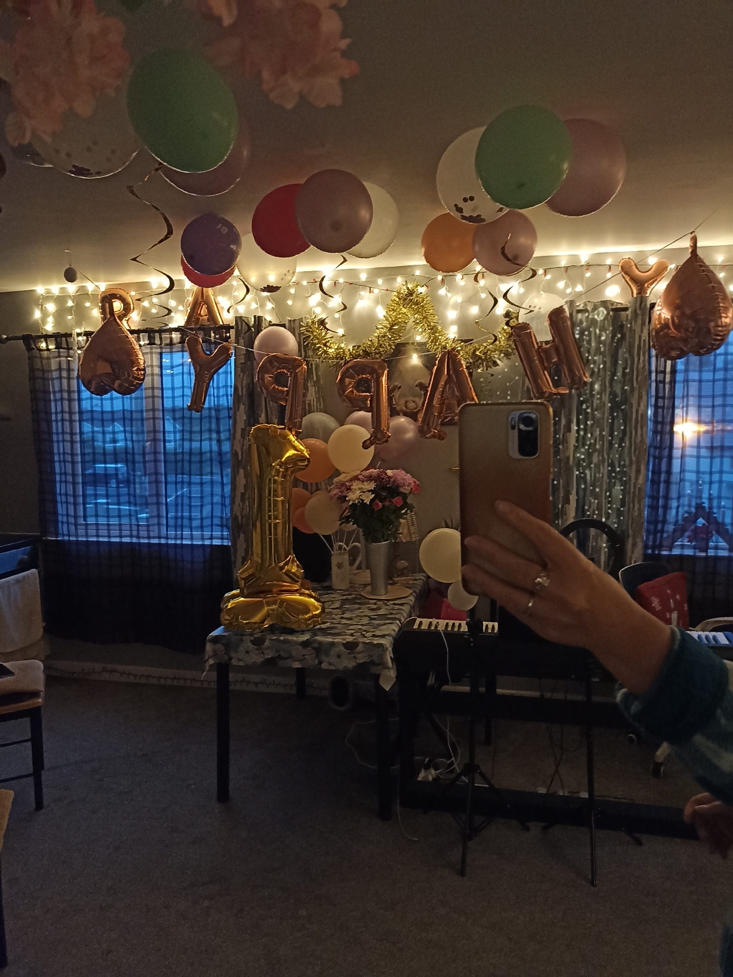 The image depicts an indoor scene decorated for a celebration, possibly a birthday. The room is adorned with numerous balloons of various colors, including green, red, pink, orange, and purple, some of which are round and others are foil letter balloons spelling out "HAPPY BIRTHDAY." There are also hanging paper decorations and string lights. In the foreground, a large golden foil balloon in the shape of the number "1" stands prominently on a table. Next to it is a bouquet of flowers in a vase. A person's hand, wearing a ring, is visible in the lower right corner, holding a smartphone, suggesting a selfie is being taken. In the background, there are curtains and a window showing a dark outdoor scene, with lights suggesting it is nighttime. A keyboard is visible on a stand in the middle ground. The overall impression is of a festive, home-based celebration. The location is identified as Annagry, Ireland.