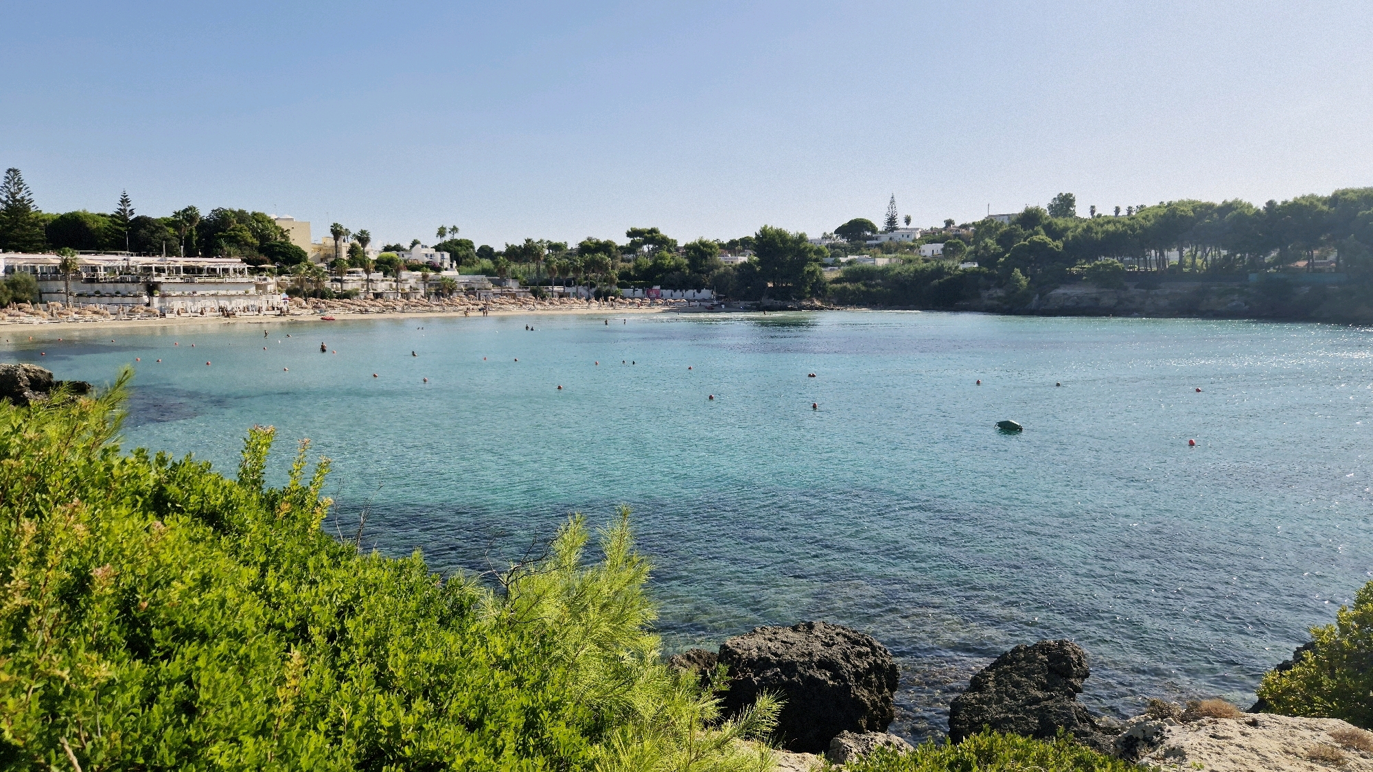 The image depicts a sunny day at a coastal location, likely in Leporano, Italy, featuring a clear turquoise sea and a sandy beach lined with sunbeds and umbrellas. Buildings with awnings and trees are visible along the shoreline, suggesting a developed tourist area. Several individuals are seen in the water, some swimming and others standing, while a small green object, possibly a raft or buoy, floats on the surface. Red buoys are also scattered across the water. The foreground is dominated by lush green foliage and dark rocks, providing a natural frame for the scene. The sky is a clear, bright blue, indicating fair weather.