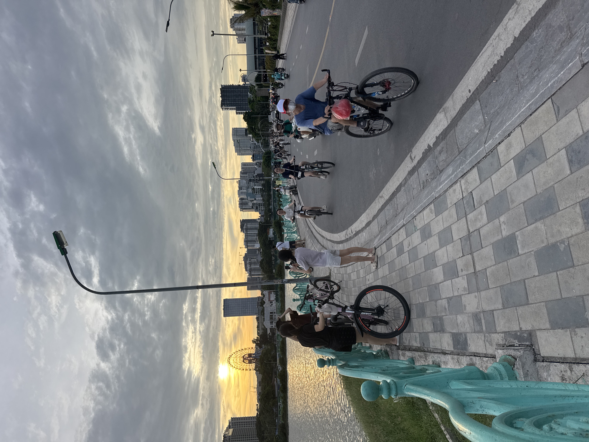 This media file captures a vibrant scene in Hanoi, Vietnam, likely during a pleasant evening. The image depicts a group of people cycling along a paved path next to a body of water. Several individuals are on bicycles, some are walking, and a couple appears to be taking a break, with one person sitting on a bike and another standing. The setting suggests a recreational area, possibly a park or promenade, with a waterfront and urban skyline in the background.
The time of day is indicated by the warm, golden light of the setting sun, casting a soft glow across the sky and water. The sky is partly cloudy, with streaks of white and grey clouds against a pale yellow and blue backdrop. This suggests fair weather, conducive to outdoor activities.
Notable details include a Ferris wheel visible in the distance, adding a landmark element to the scene. The path is paved with rectangular grey stones, and a distinctive turquoise railing lines the edge of the promenade overlooking the water. The people are dressed casually, suggesting a relaxed atmosphere. The overall mood seems peaceful and enjoyable, with a sense of community as people engage in leisure activities together. There is some text visible on the rear wheel of one bicycle, which appears to be "GIANT".