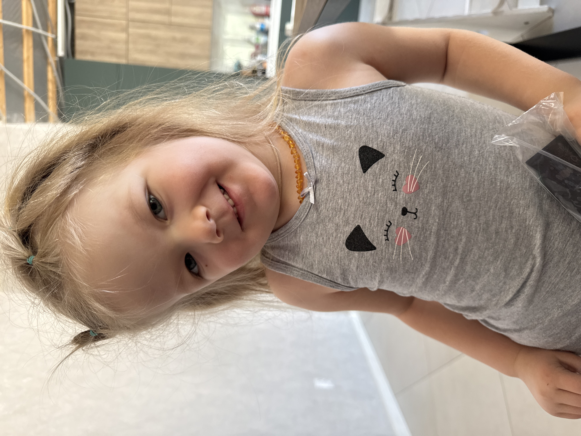 A fair-skinned child, estimated age 2-4 years, with blonde hair tied into two small pigtails with teal elastics, is captured from the waist up against an indoor background. The child wears a grey tank top featuring a black and pink cat face graphic on the chest and an amber-beaded necklace with a small silver-colored bow around the neck. The child looks directly at the camera with a slight smile. A clear plastic bag containing a dark, rectangular object is held in the child's right hand. The background, located in a residence in Gornyi (Dvurechenskoe S/P), Russia, displays a light-colored wall with a baseboard and light tiled floor to the left. To the right, a modern kitchen area with dark green and wood-grain cabinetry, a counter, and a ceiling with visible structural elements are present. The scene is illuminated by natural light.