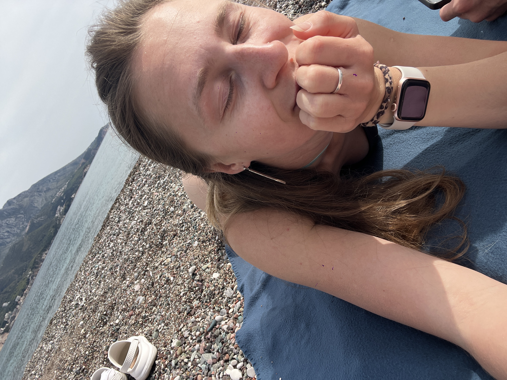 A woman is lying on a blue blanket on a pebble beach. Her eyes are closed and she is touching her face with her right hand, which has a ring on the ring finger and an Apple Watch on her wrist. She has an earring. In the background is a mountain and the sea. A pair of white shoes is partially visible on the pebble beach. A phone is visible in the upper-right corner. The location is likely the beach near Boreti, Montenegro, based on the mountainous coastal landscape and pebble beach.